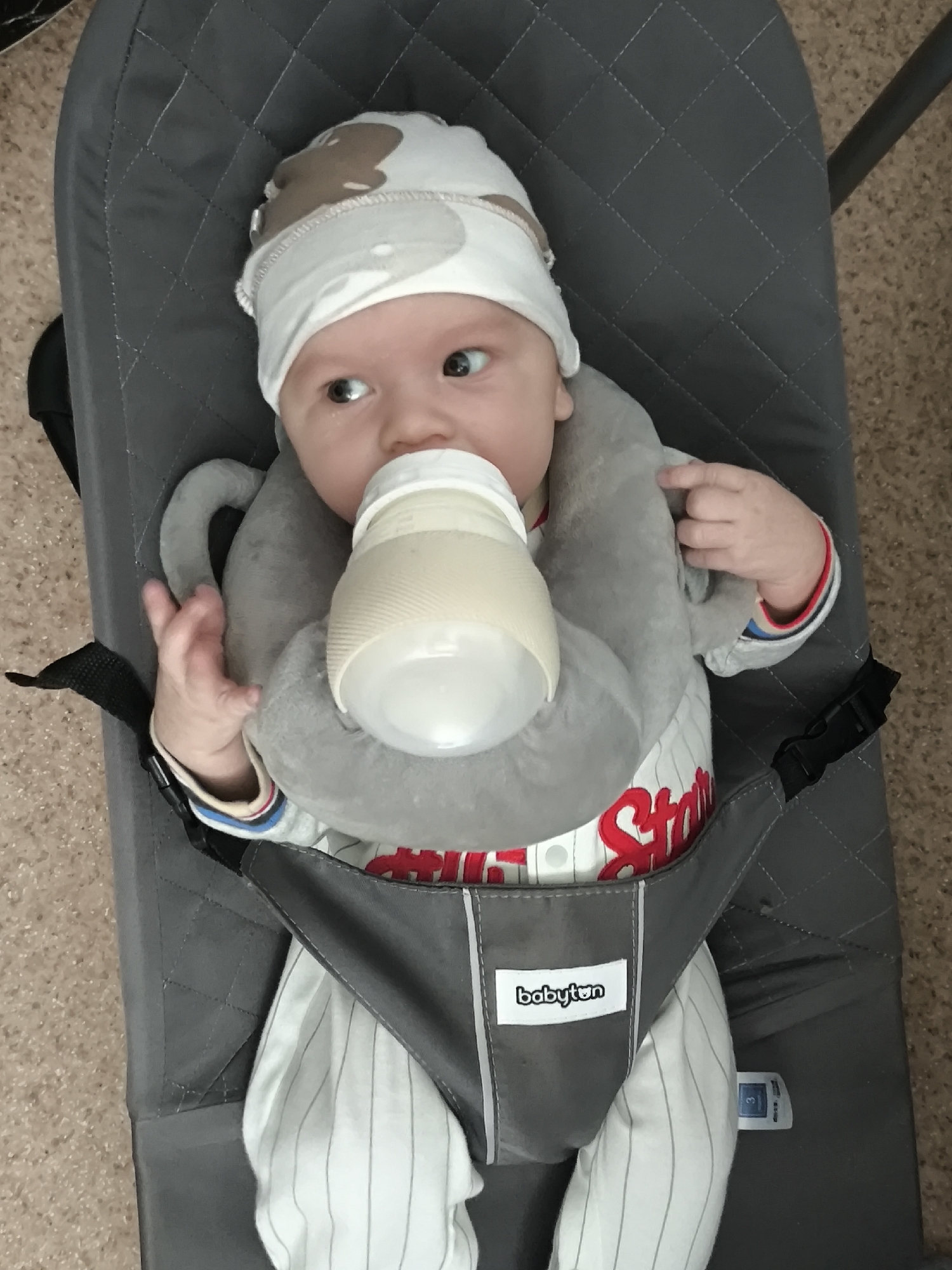 A baby is lying in a gray baby bouncer. The baby is wearing a white hat with brown spots, a white outfit with pinstripes, and a gray neck pillow. The baby is holding a bottle in its mouth. The baby bouncer has a gray harness with a "babyton" label. The background is a beige tiled floor. The location is Chaikovsky, Russia.