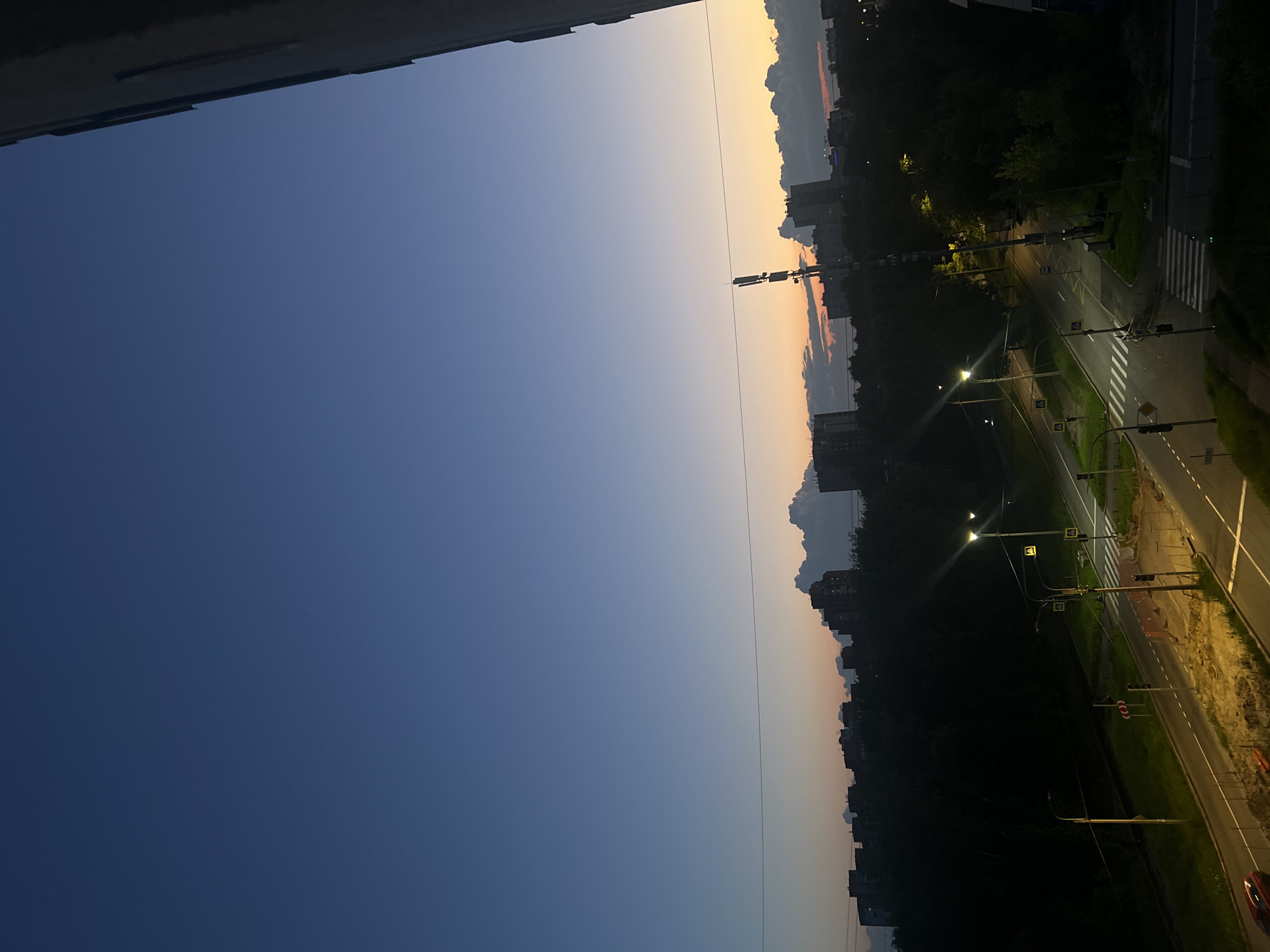 This is a high-angle, wide-shot photograph of a suburban city scene in Shushary, Russia, at dusk. The sky transitions from a deep blue at the top to lighter shades of blue, yellow, and orange near the horizon. A silhouette of apartment buildings and trees stretches across the mid-ground. A prominent cell tower stands tall amidst the buildings on the right side of the frame. In the foreground, a two-lane road with crosswalks and streetlights is visible, with a red car driving on the left lane. The scene appears to be a residential area with ongoing construction in some parts of the road, indicated by yellow barriers and mounds of dirt.
