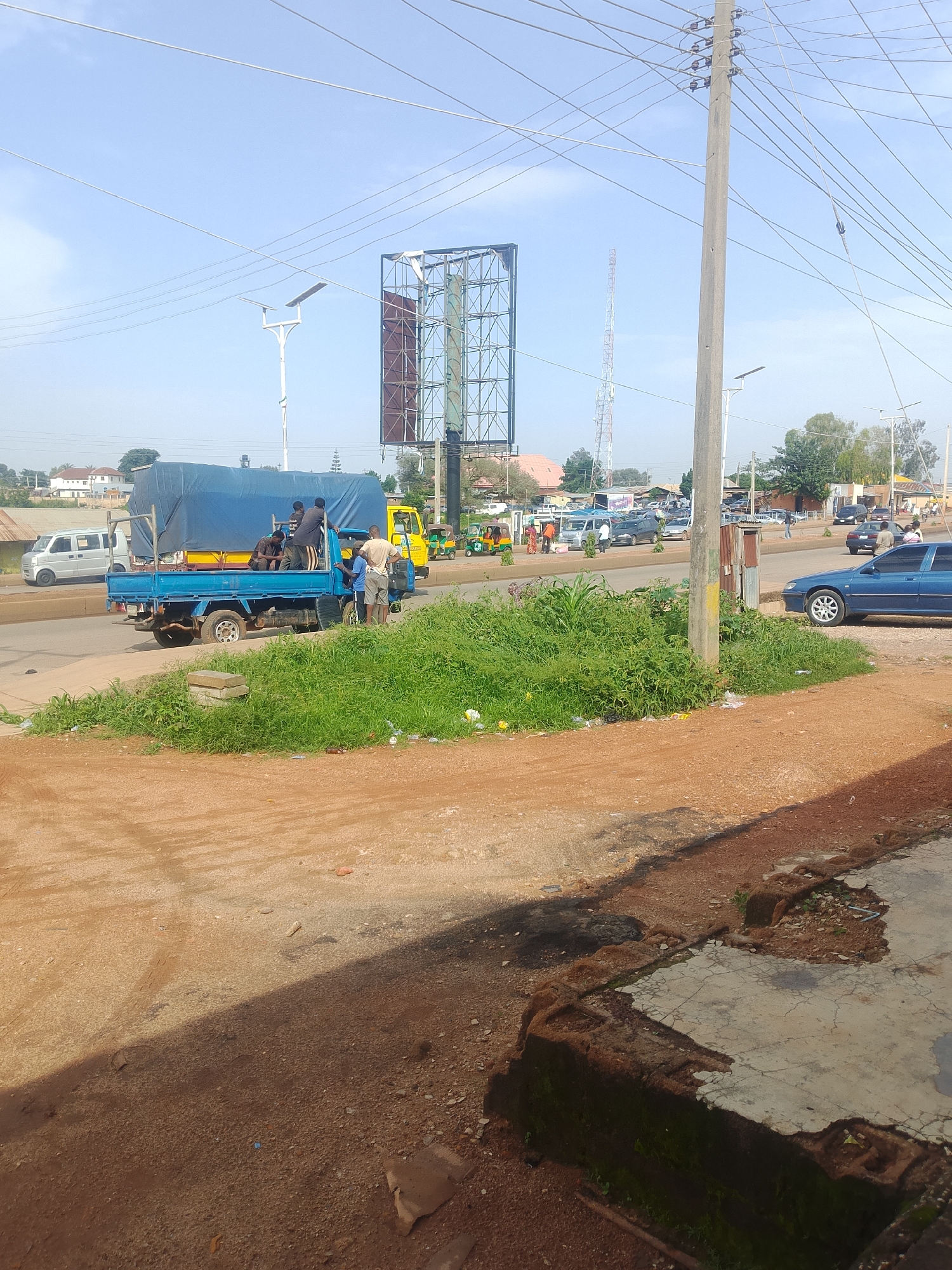 The image depicts a street scene in Bukuru, Nigeria, with a focus on a blue flatbed truck carrying people and cargo under a blue tarp. Several individuals are standing around the truck, some appearing to be passengers or workers, while others are seated in the truck bed. A yellow truck is parked behind the blue truck, and further down the road, a variety of vehicles including cars, a minivan, and a three-wheeled auto-rickshaw are visible. A large billboard structure stands prominently in the background, along with utility poles carrying numerous wires and a tall communication tower. The environment shows a paved road, a dirt area in the foreground with scattered debris and vegetation, and buildings and trees lining the street. The sky is clear and blue.