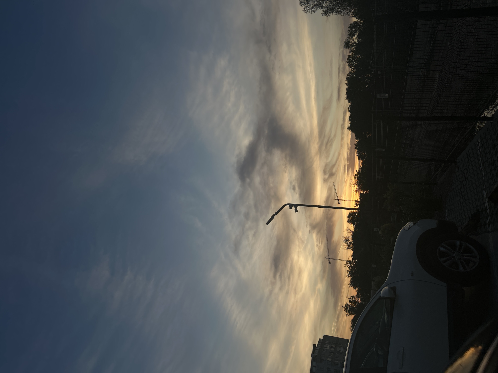 The image depicts a cityscape at sunset, likely in Plovdiv, Bulgaria, evidenced by architectural styles and local construction projects. A white car is parked on the right side of the frame, partially obscuring a metal fence and tree line. In the distance, multiple construction cranes are visible against the sky. A street lamp is positioned centrally. The sky presents a mix of blue and yellow hues, with cloud formations characteristic of sunset. A tall apartment building is present in the left corner. The lighting suggests it is near dusk.