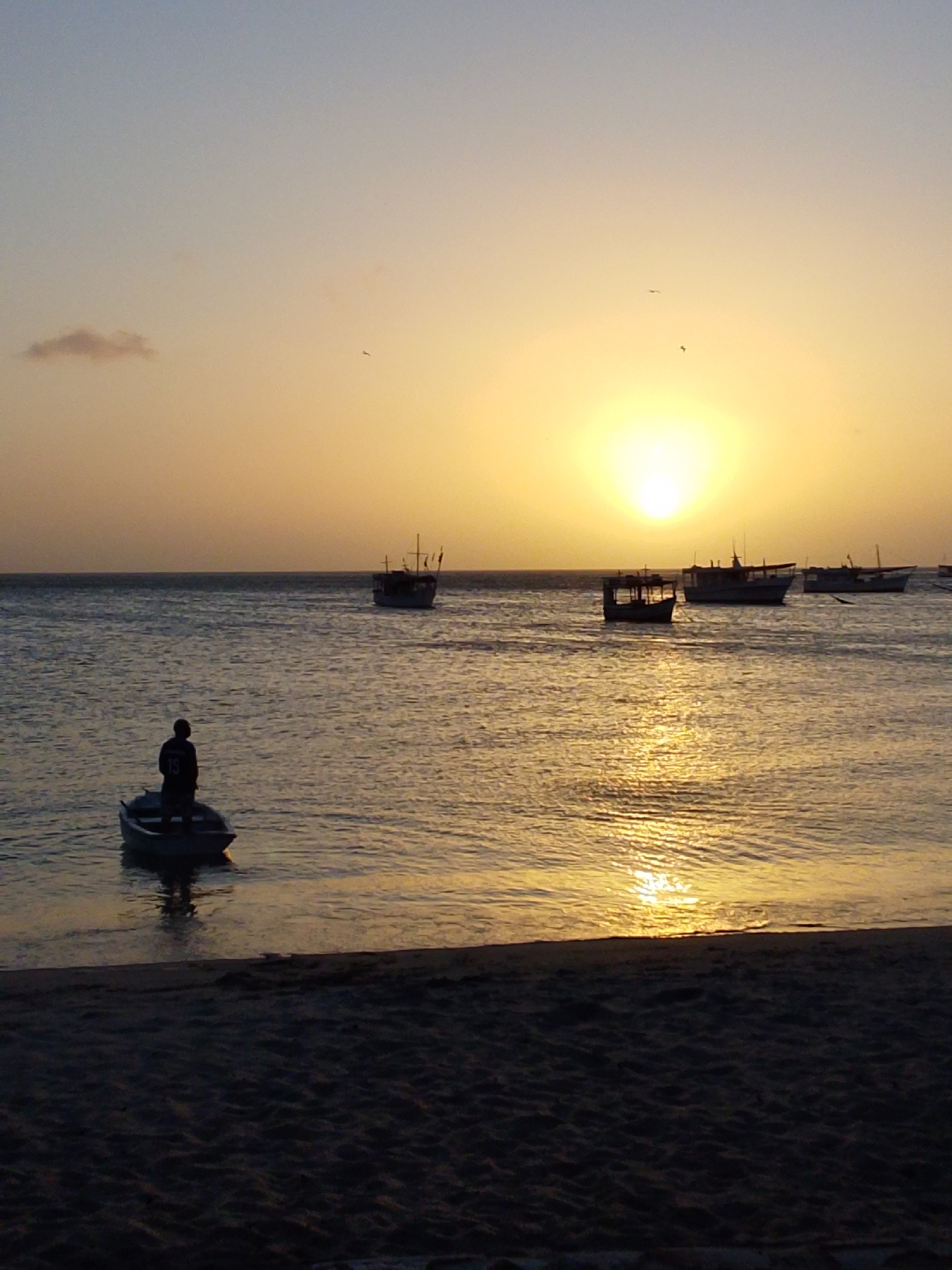 The image depicts a serene sunset over the ocean in Juan Griego, Venezuela. The sun, a bright yellow orb, is low on the horizon, casting a warm glow across the sky and water. Several boats are visible on the calm sea. In the foreground, a small rowboat with a single person standing in it is closer to the shore. Further out, larger boats are anchored, their silhouettes stark against the vibrant sky. The beach in the foreground is dark and sandy, suggesting the end of the day. The overall atmosphere is peaceful and tranquil.