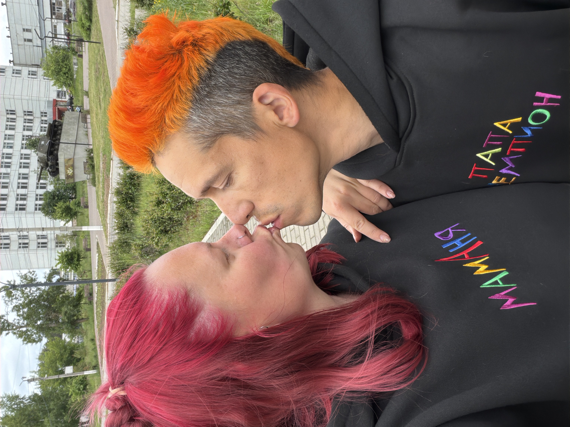 Two individuals, a woman and a man, are engaged in a kiss in an outdoor setting. The woman on the left has bright fuchsia hair, a silver nose ring, and a silver ear hoop, and wears a black hooded sweatshirt embroidered with "МАМАНЯ" (Mommy) in colorful Cyrillic letters. Her right hand, displaying light glitter nail polish, rests on the man's left shoulder. The man on the right has bright orange hair on top with gray sides and an undercut, and wears a black hooded sweatshirt embroidered with "ПАПА ЭМОЦИЙ" (Dad of Emotions) in colorful Cyrillic letters. His left hand is placed on the woman's back. Both have their eyes closed. They are standing on a paved pathway. The background consists of green grass, deciduous trees, and a multi-story light-colored building under an overcast sky. Prominently in the middle ground is a dark green ISU-152 tank monument mounted on a concrete pedestal, a landmark located in Victory Park, Chaikovsky, Russia.
