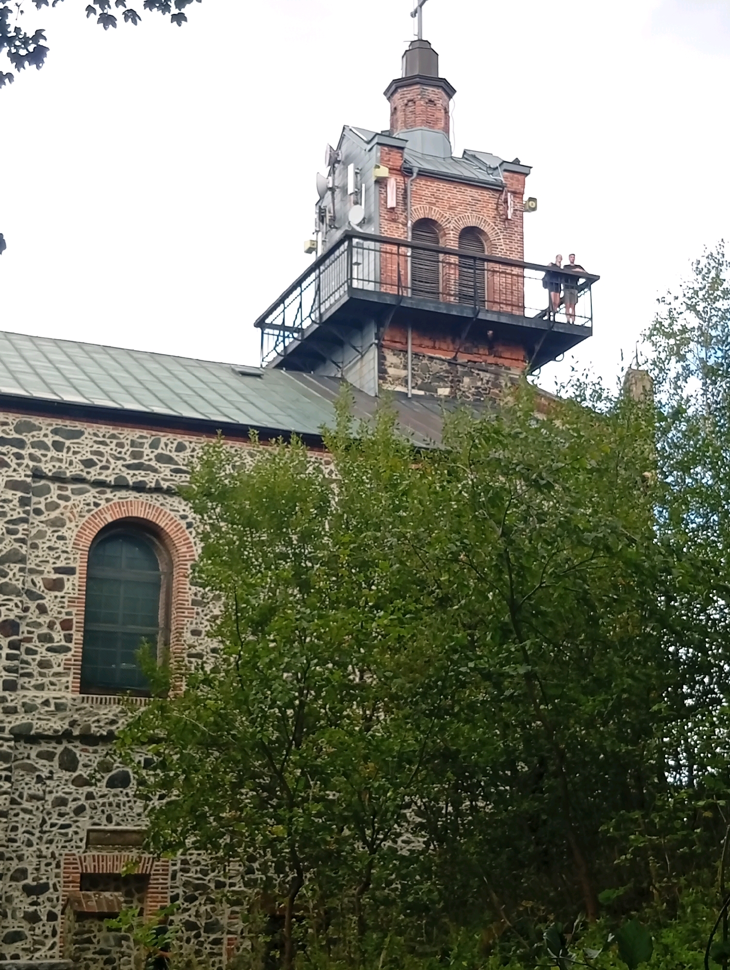 In the city of Sobótka, Poland, the image captures a close-up of a stone building with a green-tiled roof, featuring an arched window. Above the main structure, a brick tower with arched openings is visible, topped with a grey spire. A metal observation deck extends from the tower, where two individuals stand, one looking to the left and the other to the right. Lush green trees partially obscure the building and tower from view. The sky is overcast.