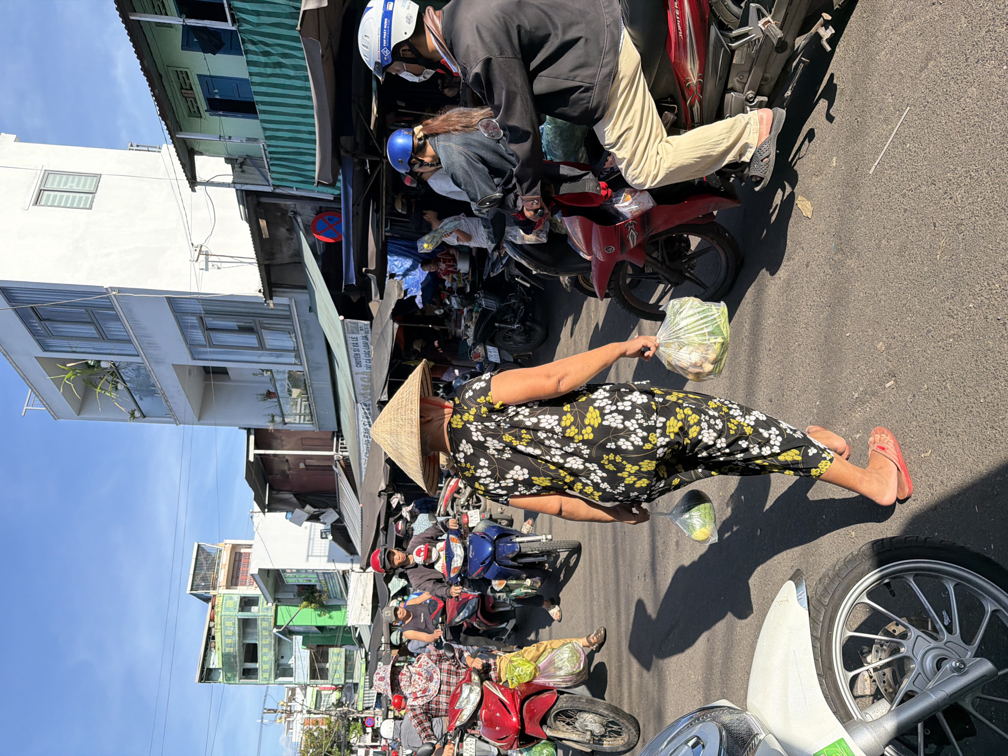 In a street scene in Nha Trang, Vietnam, a woman wearing a conical hat and a black and yellow floral print outfit walks away from the camera, carrying two plastic bags filled with produce. She is wearing red flip-flops. Several motorbikes are visible, with people riding them, suggesting a busy market or street. Some of the motorbikes are red, one is blue, and another is white. The buildings in the background are a mix of modern and older structures, with a white building on the right and a green building further down the street. The sky is clear and blue, indicating a sunny day. The scene appears to be a typical urban street in Vietnam, likely a market area with a lot of pedestrian and vehicle traffic.
