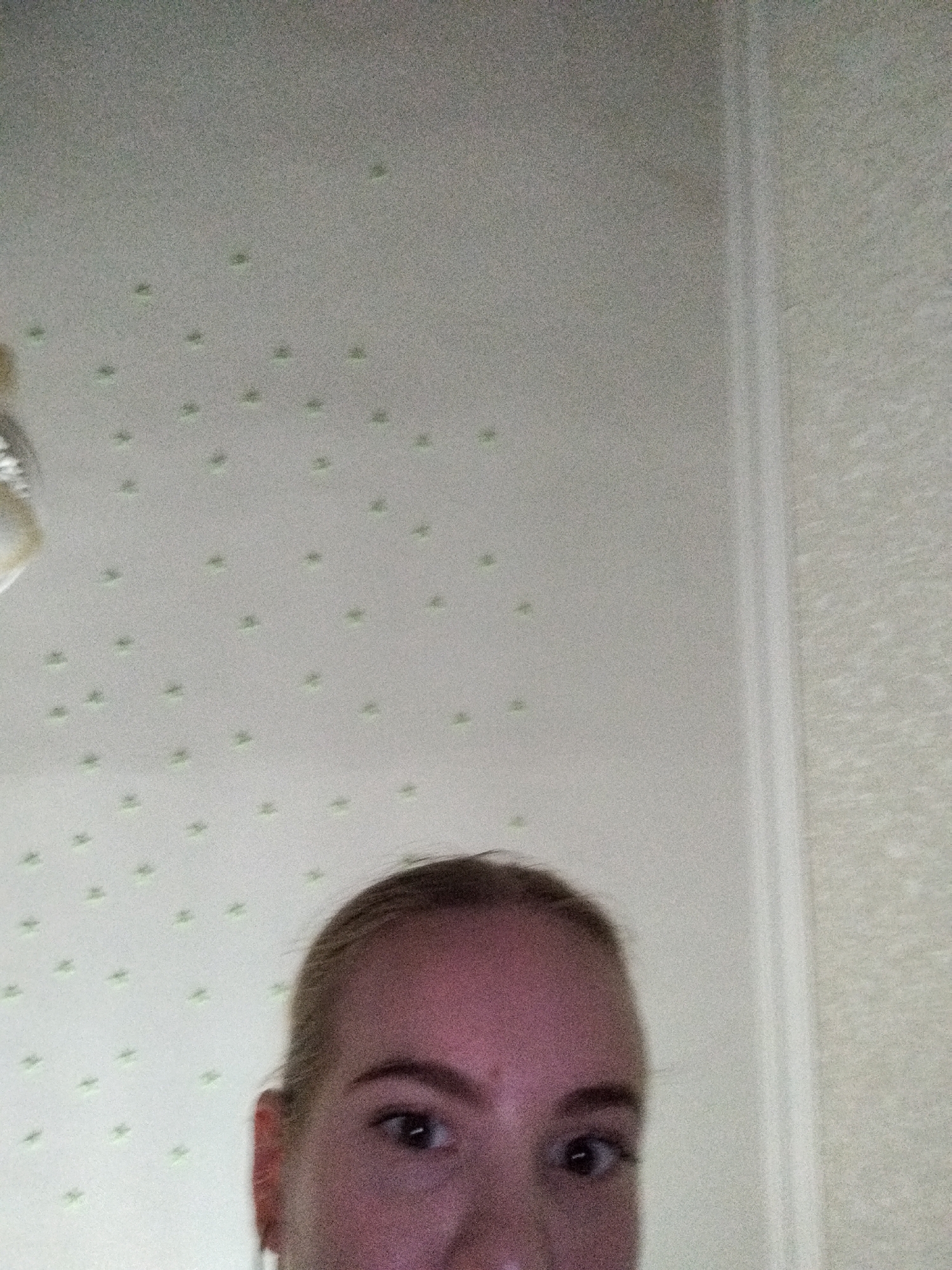 The image shows a close-up, Dutch angle shot of a young woman's face, looking up at the camera. Her left eye and brow are visible, along with a portion of her forehead and right cheek. She has fair skin and blonde hair pulled back. The background is a white ceiling with a repeating pattern of small, light green dots. The lighting is somewhat dim, casting a slight pinkish hue on her skin. The overall scene suggests an indoor setting, possibly a bedroom or living area. There is no visible text in the image.