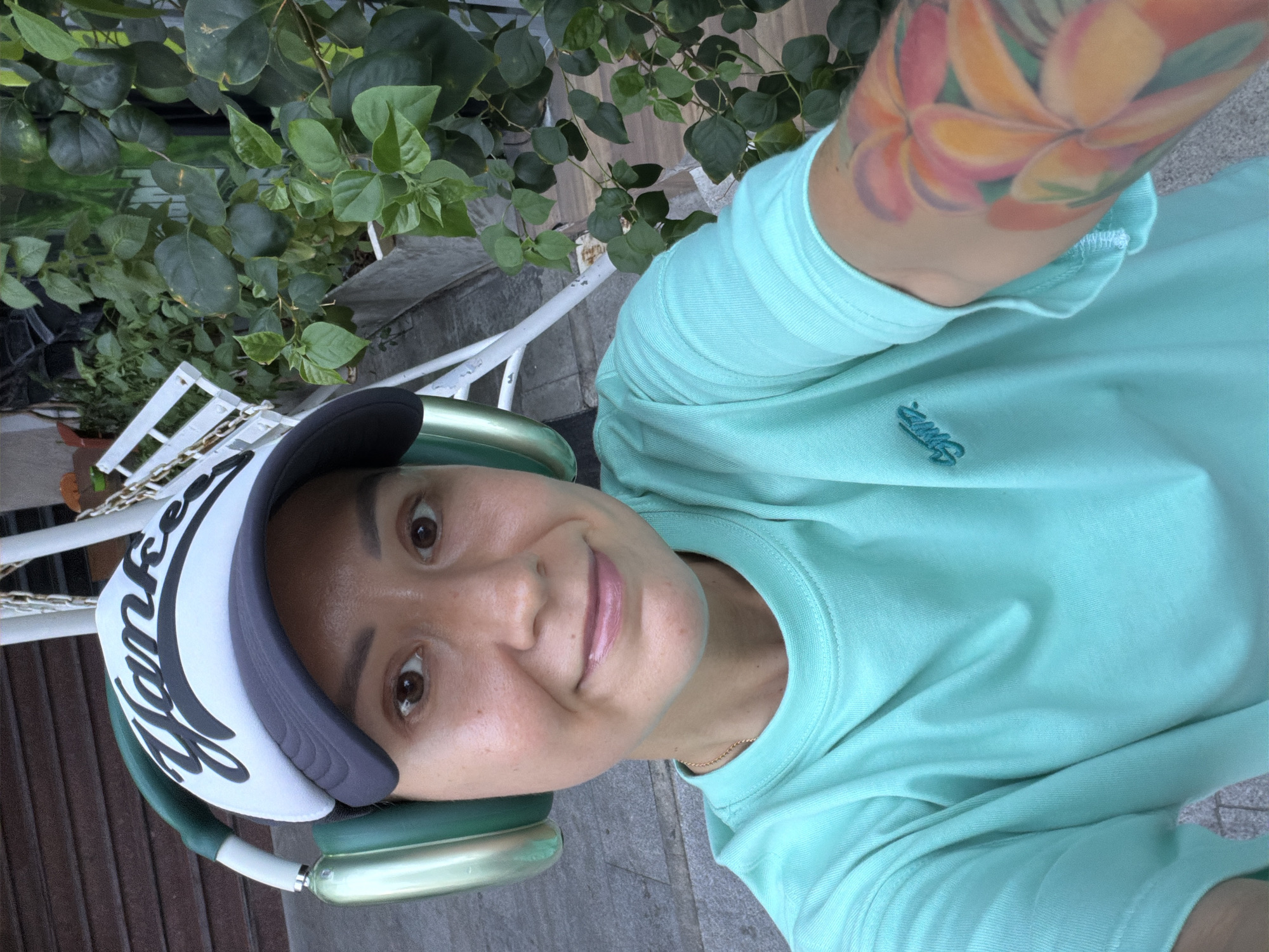 A woman wearing a turquoise t-shirt and a white baseball cap with "Yankee" written on it is taking a selfie. She is also wearing large green headphones. Her right arm, visible in the foreground, is tattooed with colorful flowers. Behind her, there is lush green foliage and part of a white structure, possibly a swing or a bench, made of metal and chains. The ground appears to be paved with grey square tiles. The scene suggests an outdoor setting, possibly in a public space like a park or a cafe with an outdoor seating area, in Nha Trang Ward, Vietnam.