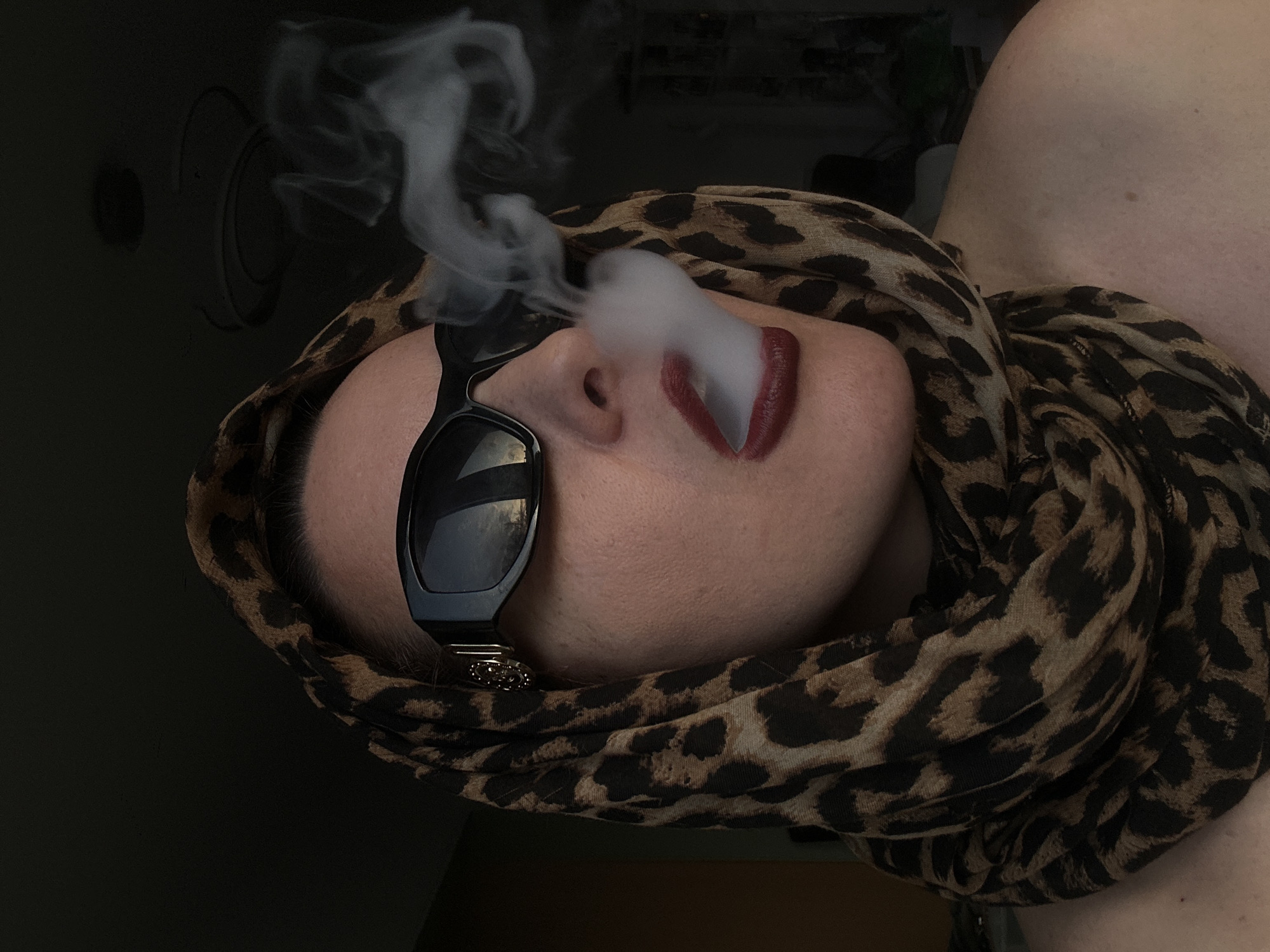 A medium shot, captured indoors under dim lighting conditions, features a person with pale skin wearing an animal print scarf, sunglasses, and dark lipstick. The person is positioned at an angle, with their face slightly tilted upwards. Plumes of smoke obscure part of the face, emanating from around the nose and mouth. The sunglasses, black and square-framed, reflect light. The scarf is draped around the head and shoulders. A dark interior, possibly a room, is visible in the background. The location is not discernible from the image.