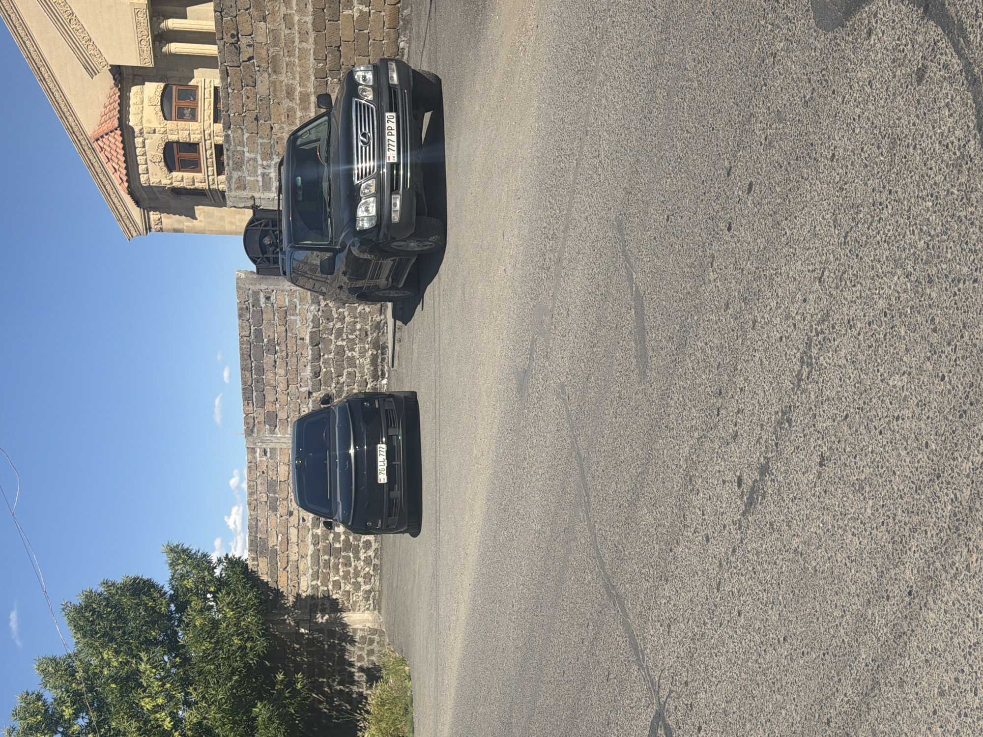 A photograph captures a street scene featuring two black Lexus LX 470 SUVs parked on a gray asphalt road. The first SUV has a license plate reading "777 PP 70," while the second displays "70 LL 777." A stone wall runs along the side of the road, adjacent to which lush green trees are visible. Above the wall, a clear blue sky is seen with scattered wisps of white clouds. In the background, a building with a textured stone facade and arched windows can be observed. The location appears to be a residential area, likely in a city or town, though its specific name remains unknown.