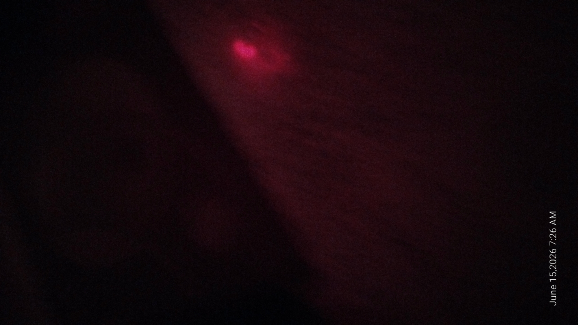 The image is a close-up, low-light photograph of a small, bright pinkish-red light source against a dark, out-of-focus background. The light source appears to be a small, irregular shape, possibly a LED or a similar point of light. The background has a reddish hue, suggesting it might be skin or a fabric material illuminated by the light. The date and time "June 15, 2026 7:26 AM" are overlaid in the bottom left corner. There is no discernible urban or environmental context within the image.