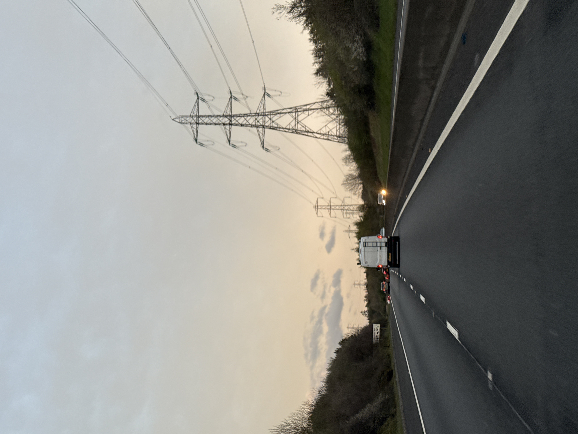 The image shows a multi-lane highway with traffic. The road is dark gray, and the sky is overcast, with a yellowish hue. Along the side of the road are trees and power lines supported by metal towers. A white van is visible ahead, with other vehicles further down the road. The location is unknown, as no specific city or identifiable features are present to determine the precise location.