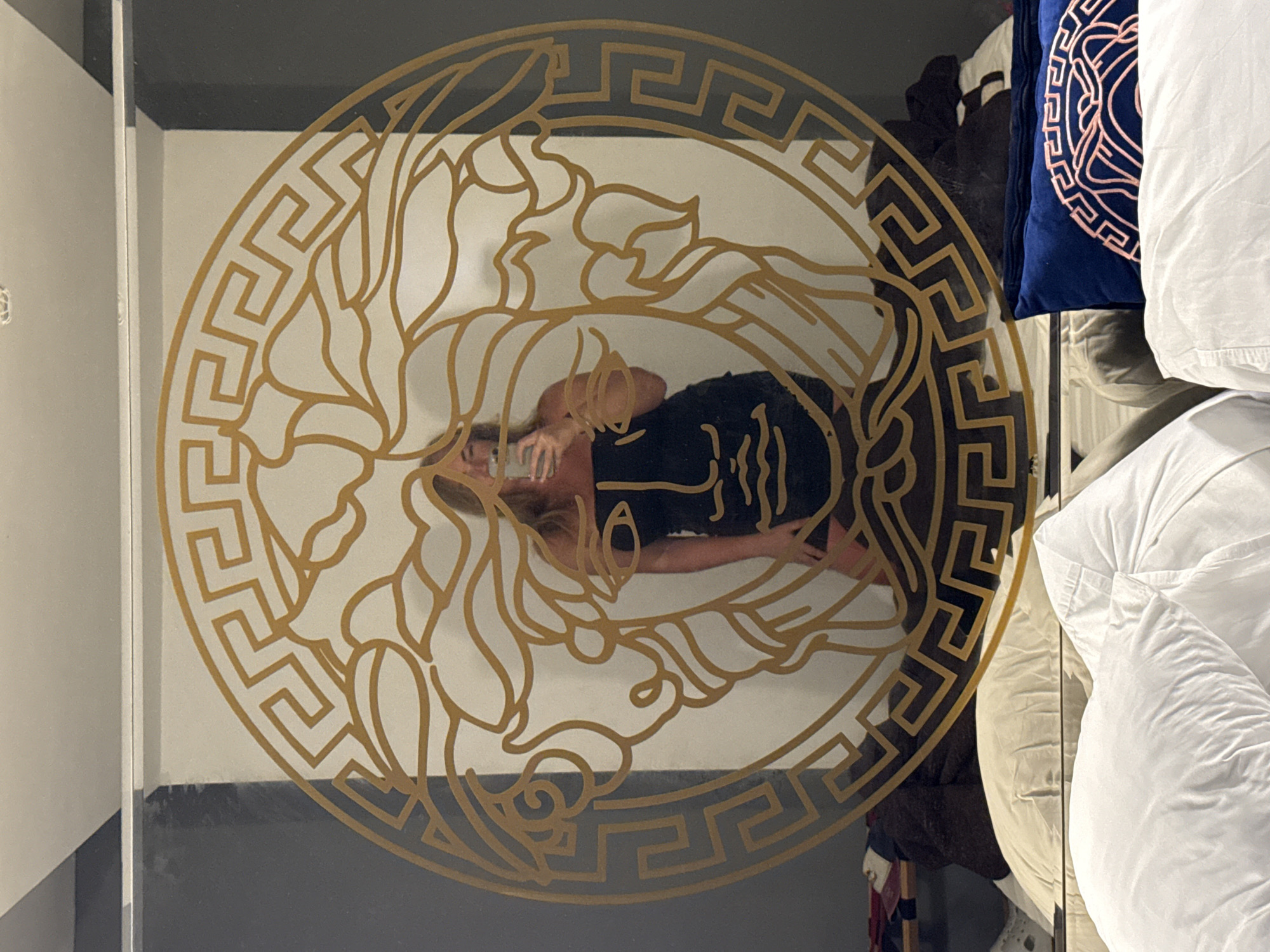 A person in a black dress is reflected in a glass object with a Versace logo design. The reflection also shows the person holding a phone. Adjacent to the glass object are bedding items including pillows and a comforter. The location is Alanya, Turkey.