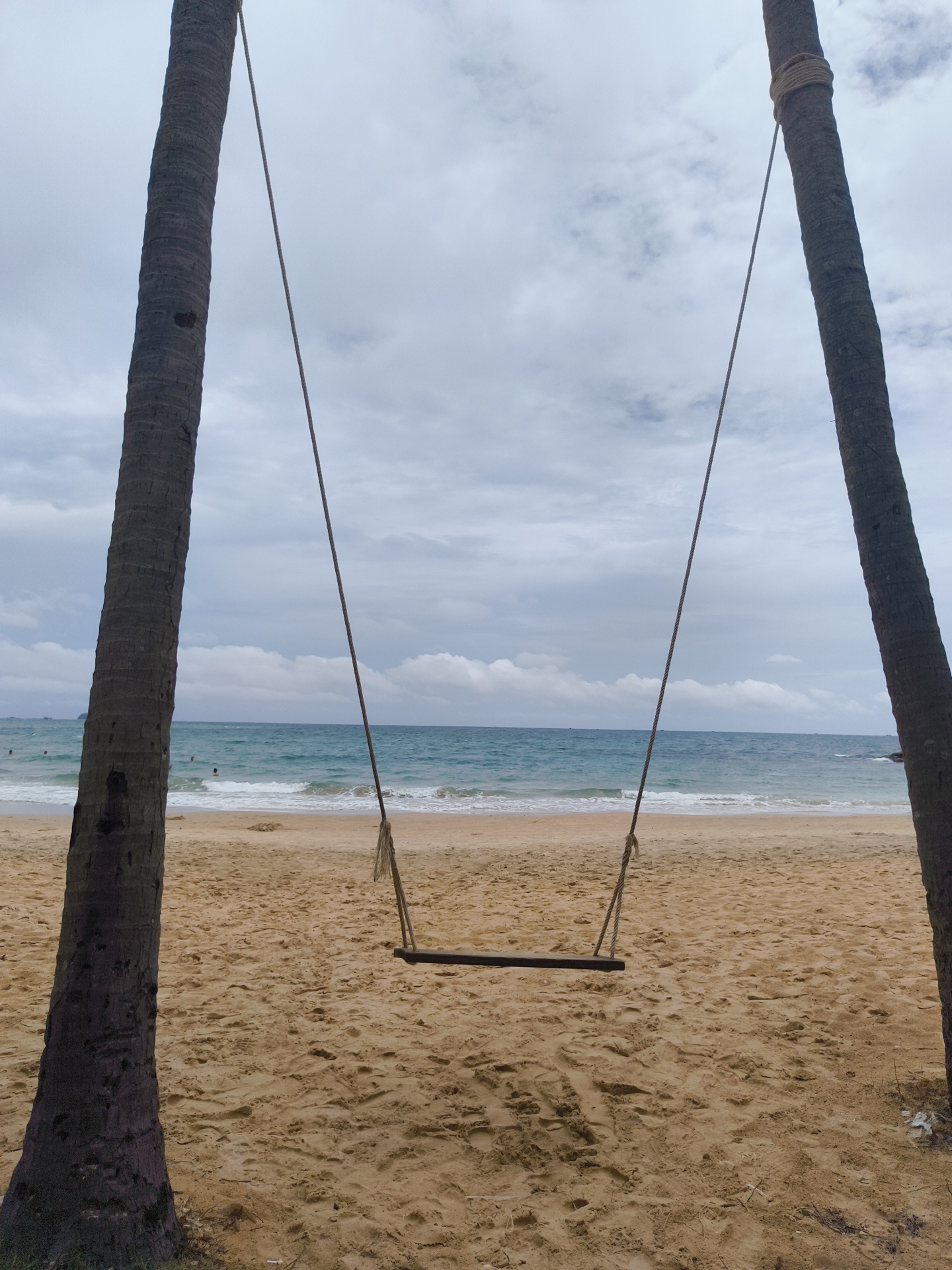 The image depicts a serene beach scene on the island of Phú Quốc, Vietnam, likely on a cloudy day. Two prominent coconut palm trees flank the foreground, their rough trunks casting shadows on the sandy beach. A simple wooden swing hangs between them, suspended by thick ropes from unseen anchor points high up on the trees. The swing is empty.
Beyond the sandy expanse, the turquoise waters of the ocean stretch towards the horizon. Gentle waves roll onto the shore, creating a foamy white line where they meet the sand. In the distance, a few small figures can be seen wading or swimming in the water, indicating the presence of other beachgoers, though the overall atmosphere is one of tranquility and solitude.
The sky above is overcast with a mixture of grey and white clouds, suggesting either a calm, diffused light or the approach of rain. The sand itself is a warm, golden hue, marked with subtle footprints and the natural undulations of the beach. The overall composition of the image creates a sense of peaceful anticipation, as if the swing is waiting for someone to enjoy the view and the gentle sea breeze. The scene is natural and uncluttered, focusing on the simple beauty of the coastal environment.