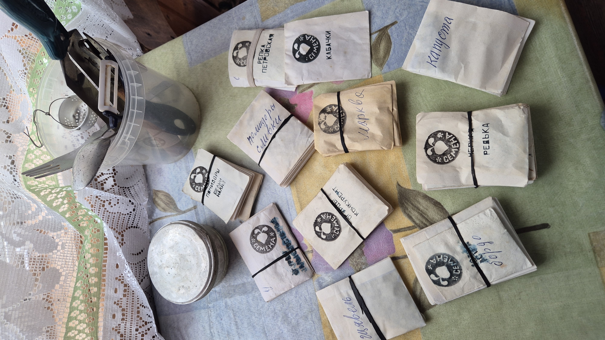 This media file depicts a collection of seed packets arranged on a colorful tablecloth, likely in a domestic setting in Машезеро, Russia. The packets are of various sizes and are held together by black rubber bands. Each packet features a circular stamp with a stylized image and the word "СЕМЕНА" (seeds). Some packets have handwritten labels in Russian indicating the type of seed, such as "РЕДЬКА" (radish), "КАБАЧКИ" (zucchini), "ПОМИДОРЫ" (tomatoes) with specific varieties like "БЕЛЫЙ НАЛИВ" and "сливки", "ОГУРЦЫ КОНКУРЕНТ" (cucumber competitor), "Капуста" (cabbage), "Марква" (likely a type of lettuce or herb), "щявель" (sorrel), and "бордо" (likely a type of beet). One packet has the handwritten word "Капуста" at the top, and another has "ДАЛЬНЕВОСТОЧНЫЕ" (Far Eastern), possibly referring to a variety.
In the background, on the left side of the image, a clear plastic container holds various kitchen utensils, including a peeler, a fork, and a spoon. A piece of white lace fabric with a green trim is also visible on the left, adding a touch of domestic detail. The lighting suggests it might be daytime, with natural light illuminating the scene. The overall impression is one of preparation for gardening or storage of seeds for future use. There are no people visible in the image.