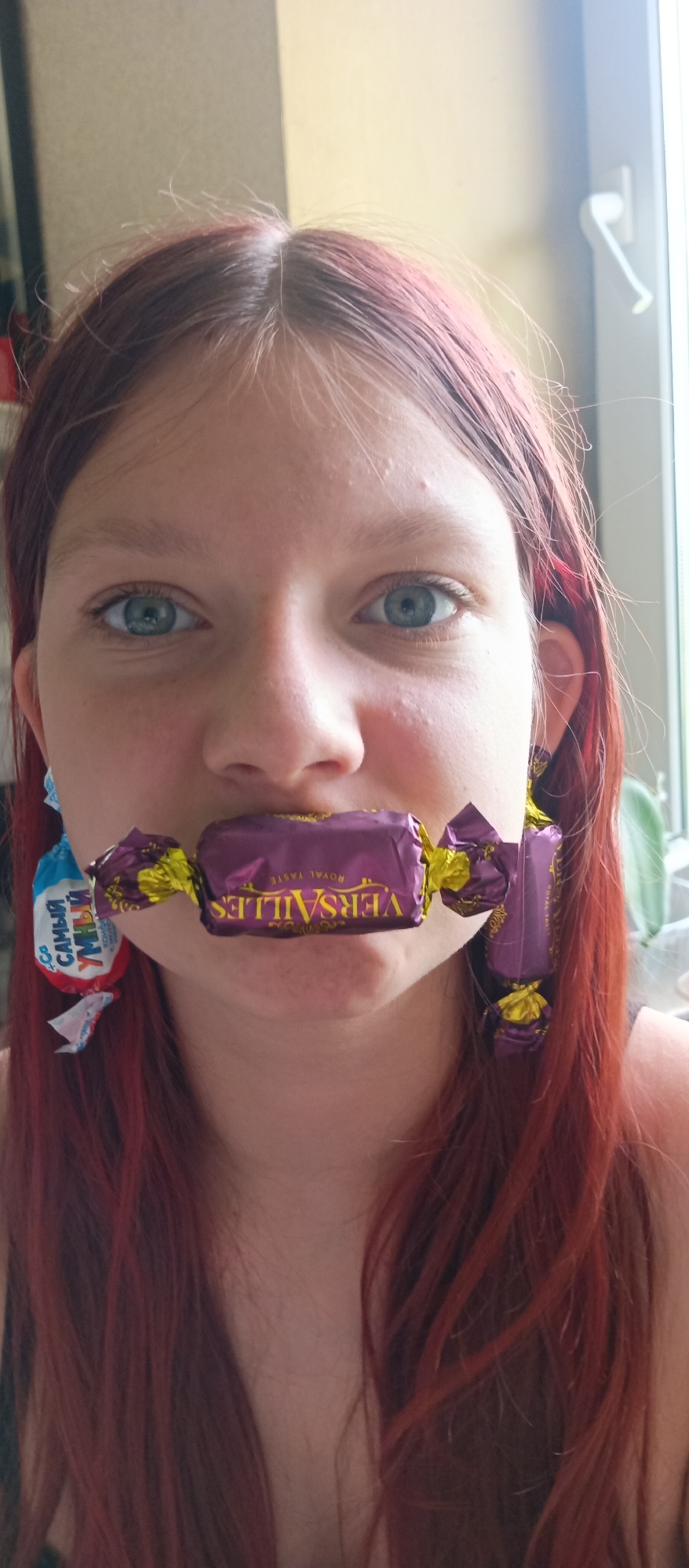 A young person with reddish-brown hair and blue eyes is positioned close to the camera. They are holding a purple candy wrapper labeled "Versailles ROYAL TASTE" in their mouth like a mouthpiece, extending across their lips. On their left ear, a blue and white candy wrapper is visible, appearing to be hanging like an earring. The background is blurred, but a window with white trim is present on the right side of the frame. The city is Unknown, Unknown.