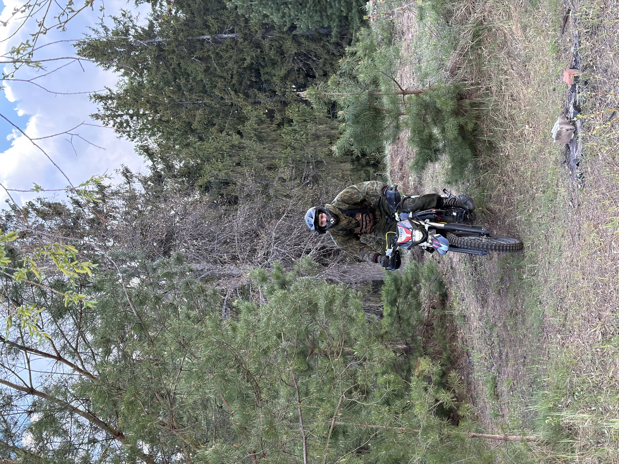 The image depicts a person wearing a helmet and camouflage clothing riding a dirt bike on a trail within a wooded area. The location appears to be Chaikovsky, Russia. The environment is characterized by pine trees and other vegetation, with a mix of clear sky and clouds visible above the trees. The dirt bike is traveling along a narrow path. Debris such as a white bag and a red object are visible on the ground to the right of the image.