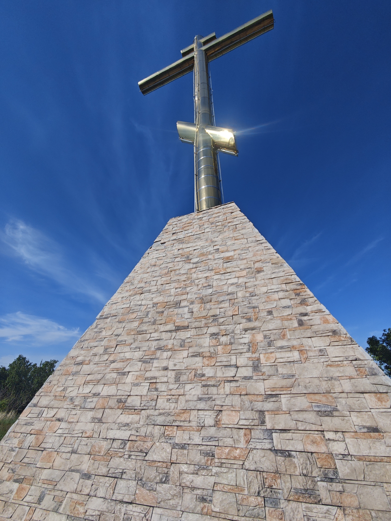The image shows a large, golden Orthodox cross standing atop a stone pyramid in Volzhskiy, Russia. The cross is made of polished metal, reflecting the sunlight. The pyramid is constructed of irregular stone blocks in various shades of tan and brown. The sky is bright blue with wispy white clouds. Trees are visible in the lower corners of the image.