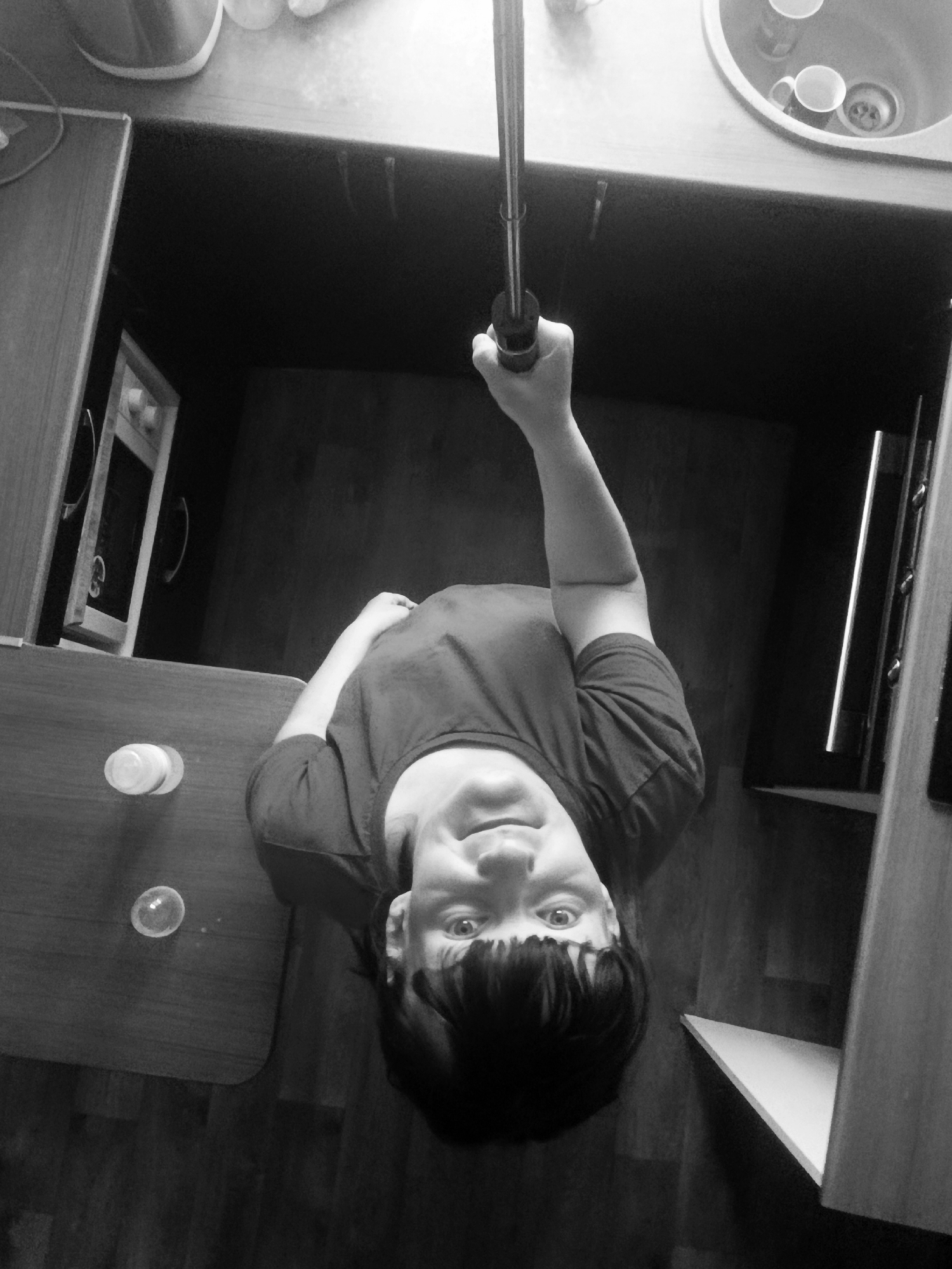 A black and white, high-angle selfie depicts a person holding a selfie stick. The person has dark hair with bangs, and is wearing a dark shirt. The background shows a kitchen environment. A sink with cups inside is visible in the upper right. Cabinets are visible on the left and right sides, and a microwave is set within one of them. Two small glass containers sit on a table to the left of the person. The location is not evident based solely on visual elements.