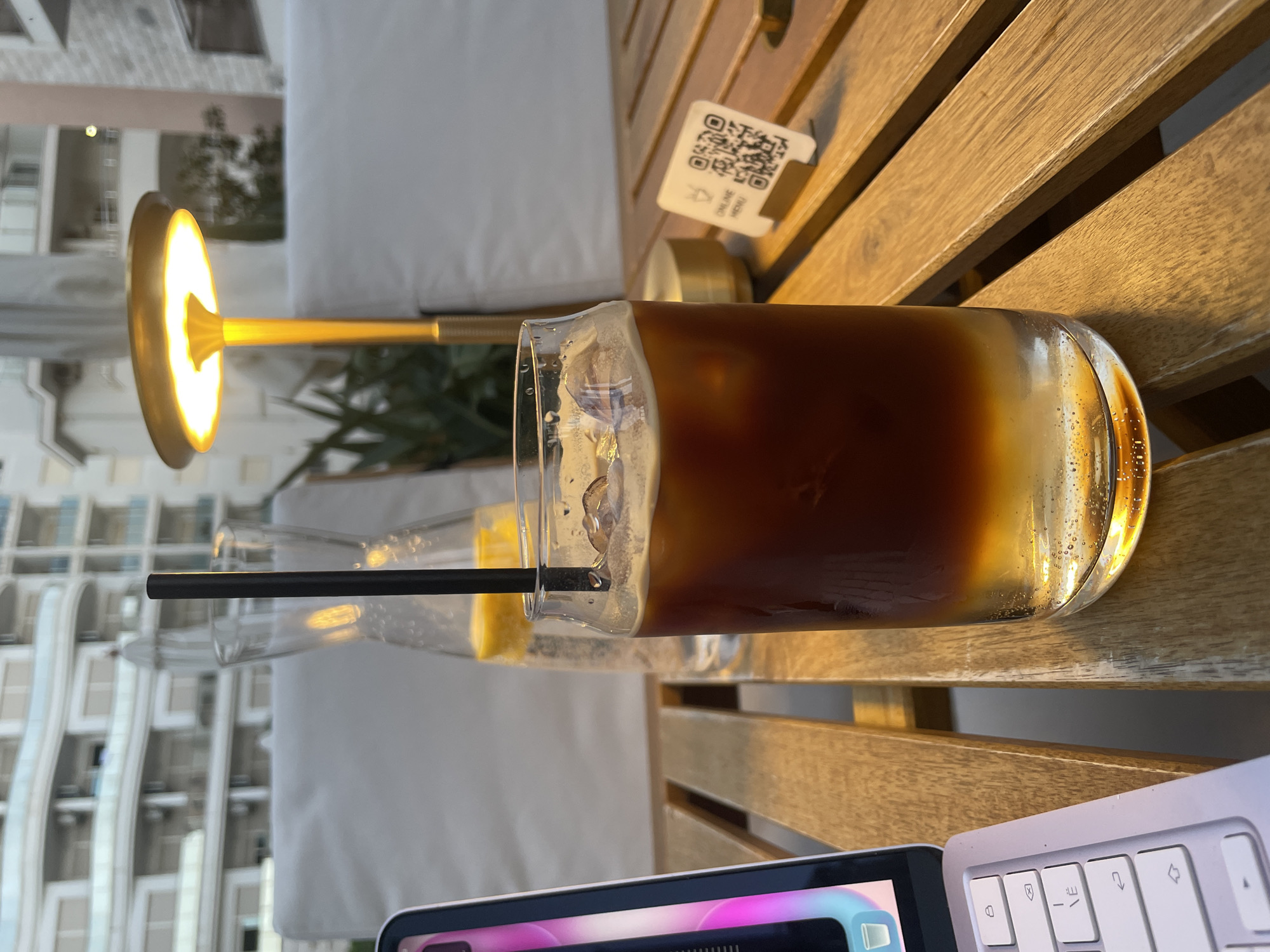 A still-life photograph taken on a wooden table shows a glass of iced coffee with a black straw, a glass with a yellow liquid inside, a small lamp, a QR code menu, and a laptop. The photo appears to be taken outdoors during the day as buildings are visible in the background. The location is potentially Boreti, Montenegro, based on the prompt.