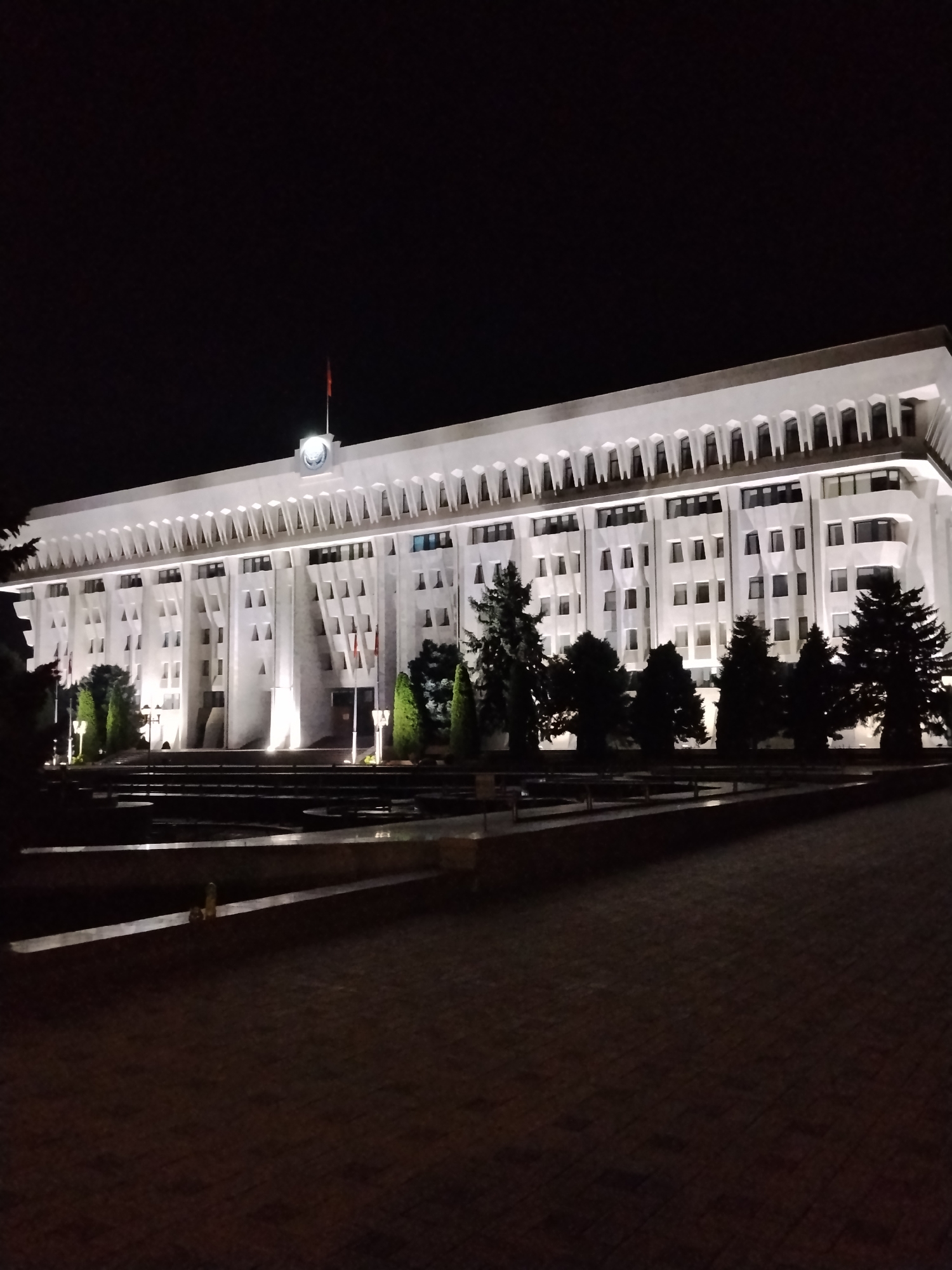 The image depicts the Government House in Bishkek, Kyrgyzstan, at night. The building is a large, white, multi-story structure with numerous windows illuminated from within. It is characterized by a grand, imposing facade featuring repeating architectural elements, possibly columns or arches, along the upper levels. A flag, red with a yellow emblem, flies from a pole on the roof, illuminated by a spotlight. In front of the building, a dark expanse is visible, likely a plaza or courtyard, with what appears to be a water feature or reflecting pool, also dimly lit. Evergreen trees line the front of the building, silhouetted against its illuminated facade. The overall atmosphere is formal and slightly dramatic due to the nighttime lighting.