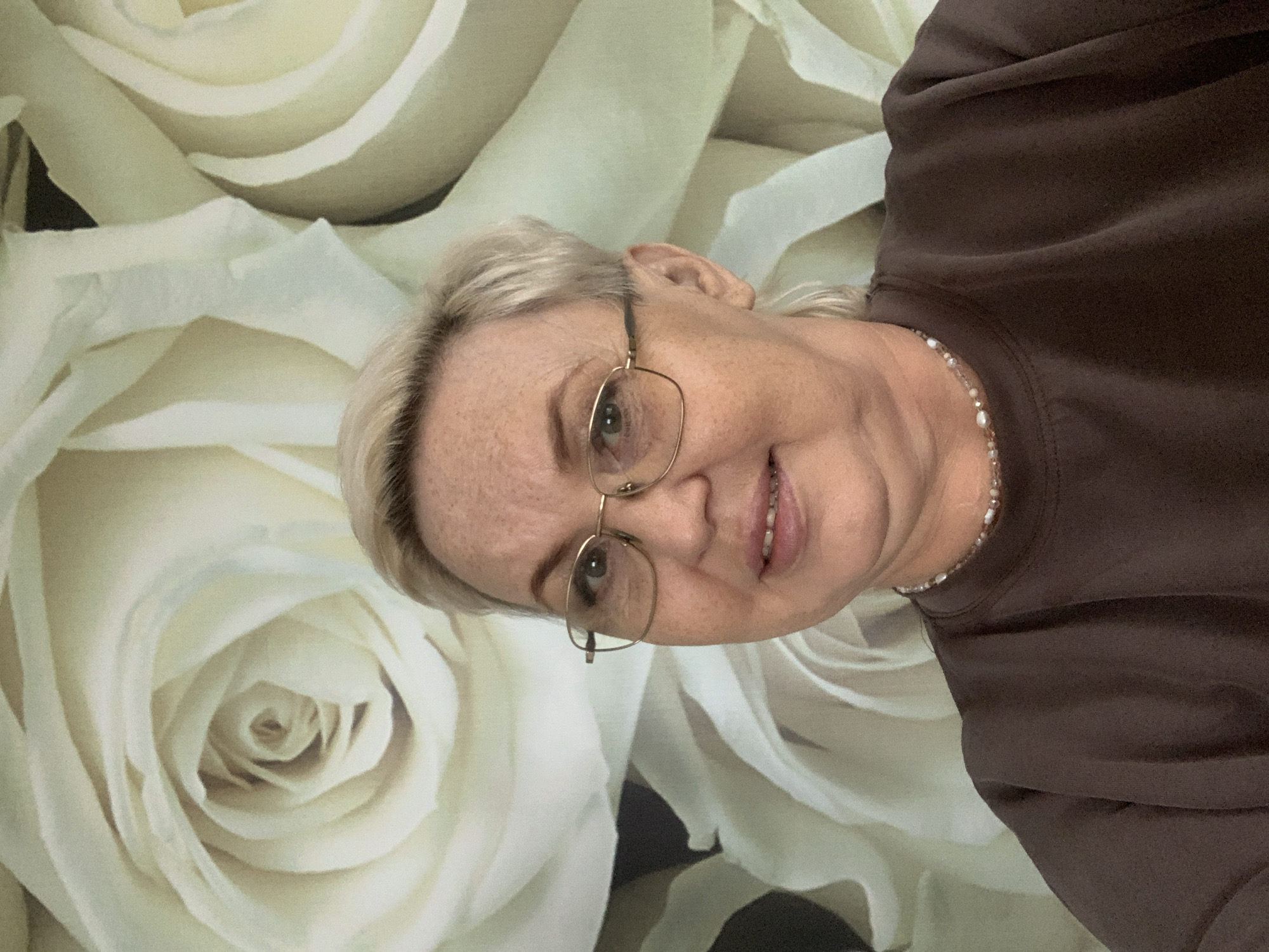 A woman with short blonde hair and glasses is seen in a close-up headshot. She is wearing a dark brown shirt and a beaded necklace. The background is a wall covered in a pattern of large, white roses. The image does not provide enough information to determine if the photo was taken in Chaikovsky, Russia.