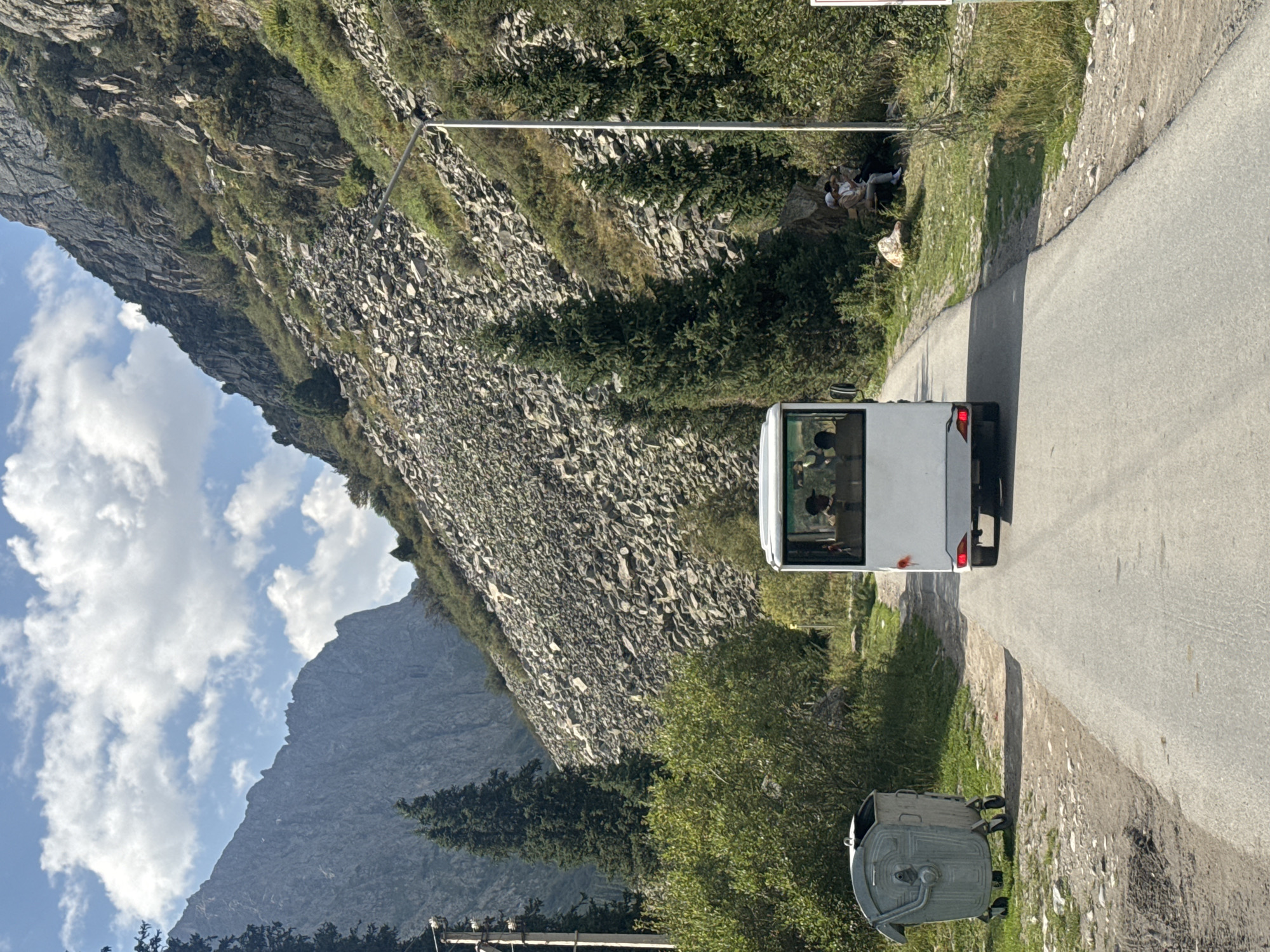 In an outdoor scene at an undisclosed location in Kyrgyzstan, a white bus travels down an asphalt road flanked by rocky slopes and lush green vegetation. The bus, with several passengers visible through its windows, is the central focus. To the left of the road sits a large, grey wheeled garbage bin. On the right side of the road, a group of individuals are seated on a bench near a lamppost, with more people visible in the background. The rugged mountainous terrain with scattered rocks and trees suggests a natural, possibly remote, environment. The sky is partly cloudy, with distinct white clouds against a blue backdrop.