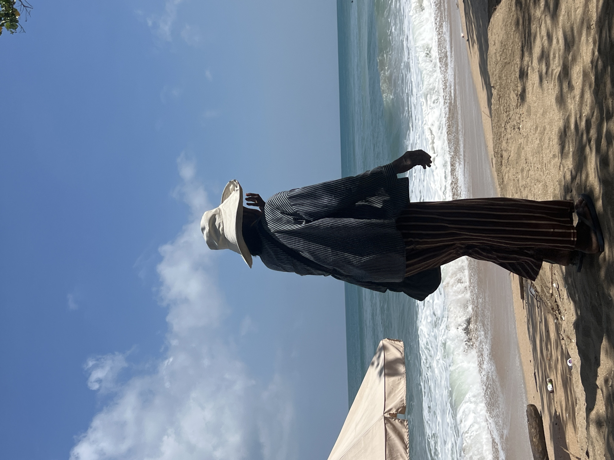 A person stands on a sandy beach in Kuta, Indonesia, facing the ocean. The person is wearing a wide-brimmed hat, a striped jacket, and striped pants. They have a bag slung over their shoulder. The ocean is calm with gentle waves breaking on the shore. An umbrella is visible to the left of the frame. The sky is blue with scattered clouds. The beach appears to have some scattered debris.