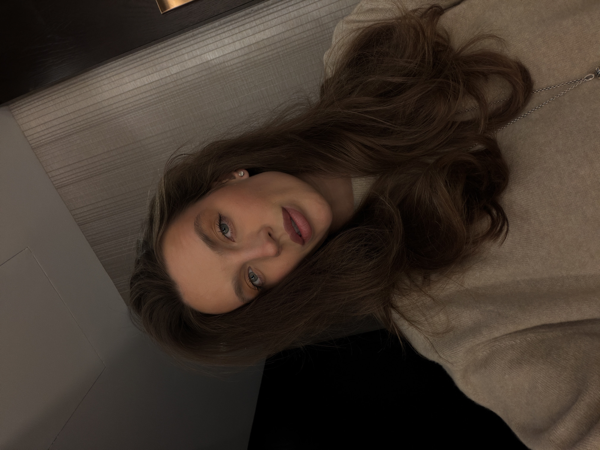 A young woman with long brown hair is the focal point of the image. She is wearing a beige sweater and a small earring is visible on her left ear. A silver chain is draped over her shoulder and sweater. Her blue eyes are visible and she has pink lips. She appears to be lying on her side. The background is a combination of neutral grey and black, with a hint of wood texture visible at the top. The scene is dimly lit. There is no indication of Budva, Montenegro, in this image.