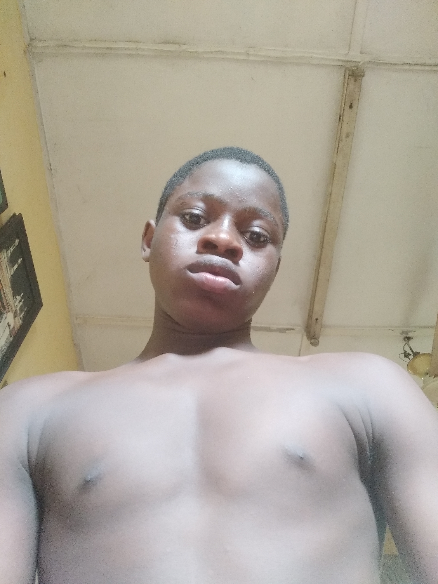 A low-angle shot shows a shirtless young Black man with a serious expression. He has short, dark hair and appears to be looking directly at the camera. The background consists of a plain, off-white ceiling with visible beams or supports. In the left background, part of a framed picture on a yellow wall is visible. The environment suggests an indoor setting, possibly a home. The location is Shangisha, Nigeria.