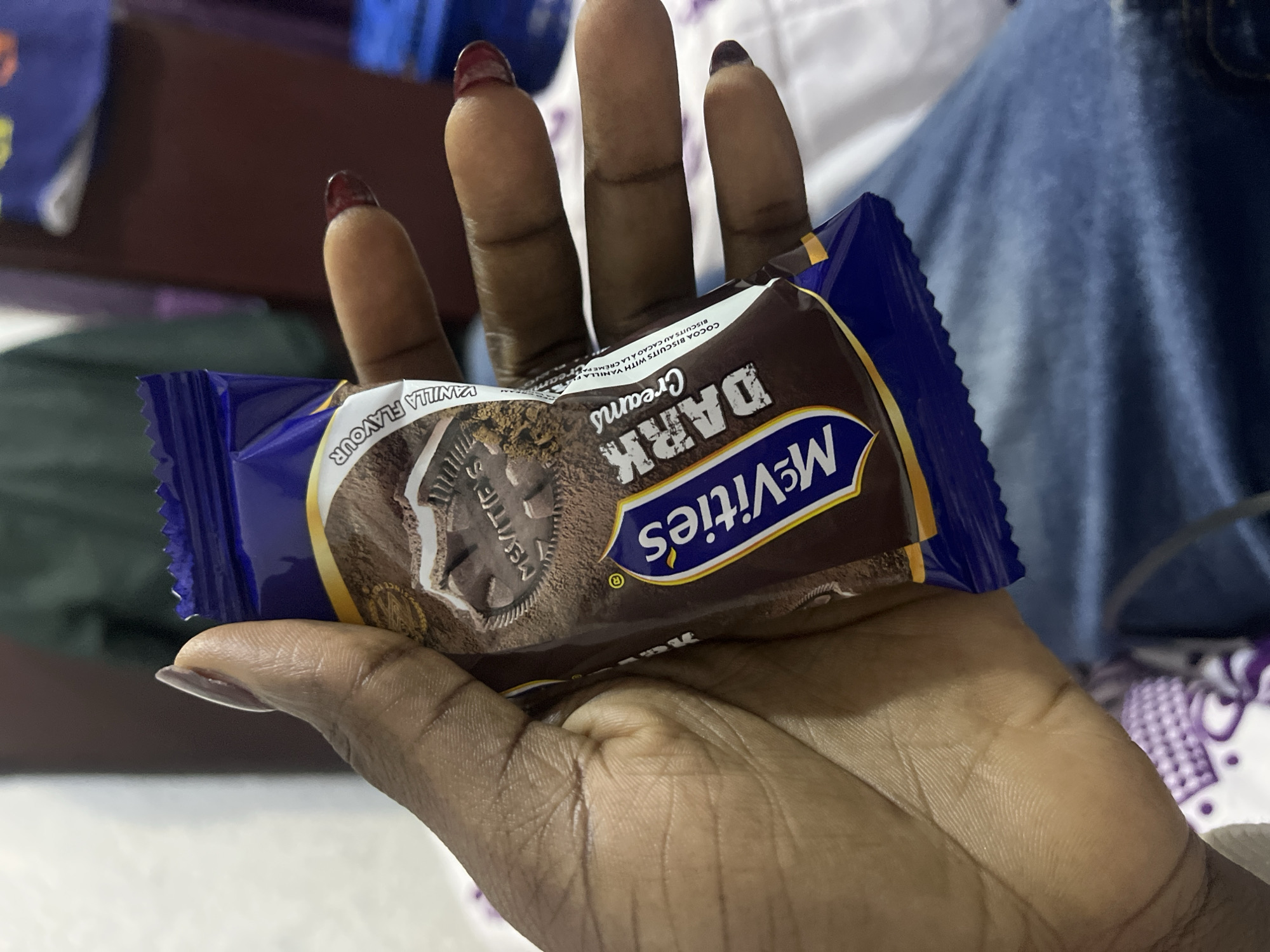 A left human hand with dark brown skin tone is depicted holding a single, unopened package of McVitie's Dark Creams biscuits. The hand displays manicured fingernails; the thumb has light purple polish, while the other four visible fingers have dark red polish. The snack packaging is dark blue and brown, featuring the 'McVitie's' logo prominently in blue and yellow. Text visible on the packaging includes 'DARK Creams,' 'VANILLA FLAVOUR,' and 'COCOA BISCUITS WITH VANILLA CREAM FILLING.' The background indicates an indoor setting. Below the hand, blue denim fabric is visible, along with white fabric patterned with purple designs, likely a bedsheet or blanket. Further back, blurred elements include dark red, dark green, and light white areas, suggesting furniture or walls. No discernible visual elements within the image provide specific indicators of the city of Abuja, Nigeria, or any particular geographic location.