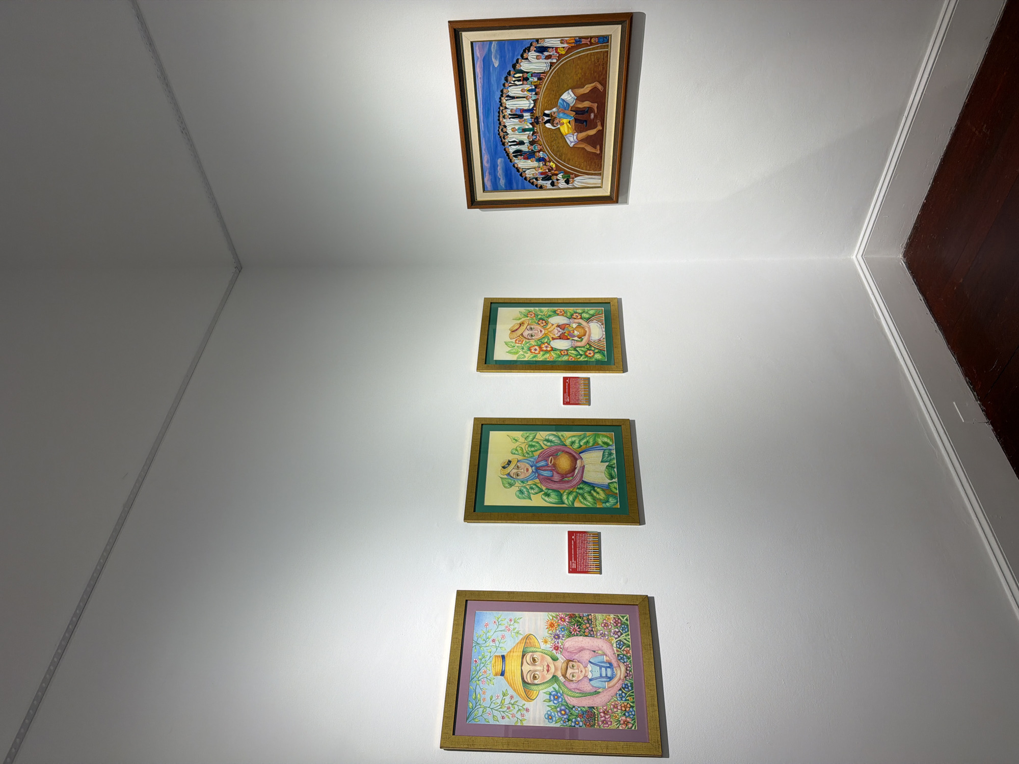 The image shows a corner of a gallery wall in La Orotava, Spain, displaying four framed paintings. The walls are painted white, and the floor is dark wood.
From left to right:
The first painting features a woman wearing a wide-brimmed straw hat and a pink dress, holding a baby. She is surrounded by a vibrant floral background with trees. The frame is gold. Two small red placards are affixed to the wall to the right of this painting.
The second painting depicts a woman with a headscarf, holding an earthenware pot. She is standing in front of a background of green foliage and orange flowers. The frame is green and gold. A red placard is on the wall to the right of this painting.
The third painting shows a woman in a yellow headscarf and a white dress, holding an earthenware pot. She is positioned in front of green foliage with orange flowers. The frame is green and gold. This painting is smaller than the first two.
The fourth painting, in the far right corner, is larger and shows a scene of a group of people gathered in a semi-circle, watching two figures wrestling in the center. The figures in the wrestling ring are wearing blue and yellow singlets. The frame is wood with a darker wood inner border.