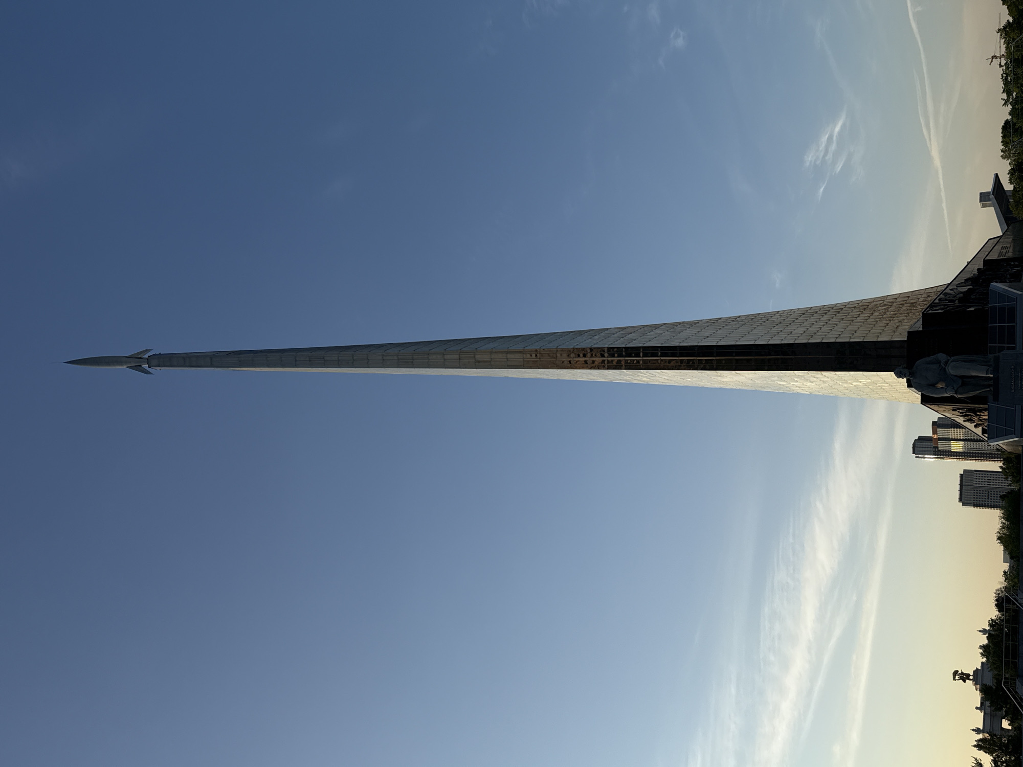 The image shows the Monument to the Conquerors of Space in Moscow, Russia. The monument is a tall, gleaming obelisk, topped with a rocket. At the base of the monument is a detailed relief sculpture depicting figures related to the space program. The monument is set against a clear blue sky, with buildings and trees visible in the background.