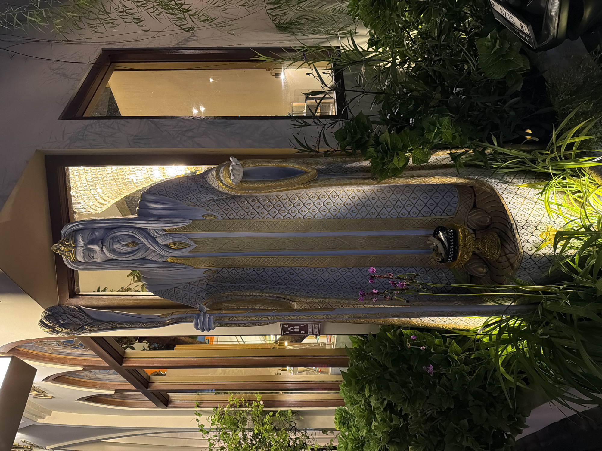 This media file features a large, ornate statue of a figure with distinct facial features, long hair, and wearing an elaborate robe adorned with a diamond pattern and gold accents. The statue is positioned in front of a building with a large window reflecting ambient light, suggesting an indoor space with chandeliers. Green foliage and plants surround the statue, creating a somewhat overgrown and naturalistic setting. The statue appears to be holding a staff or scepter.
The image is captured at night, indicated by the artificial lighting illuminating the statue and the interior of the building. The overall mood is somewhat mysterious and grand due to the scale of the statue and its decorative elements.
While the exact activity is not clear, the presentation suggests a display or an artistic installation. The location context points to Ubud District, Indonesia, which is known for its cultural and artistic attractions. There is a small sign visible in front of the statue with some text, but it is too small and blurry to be read.
