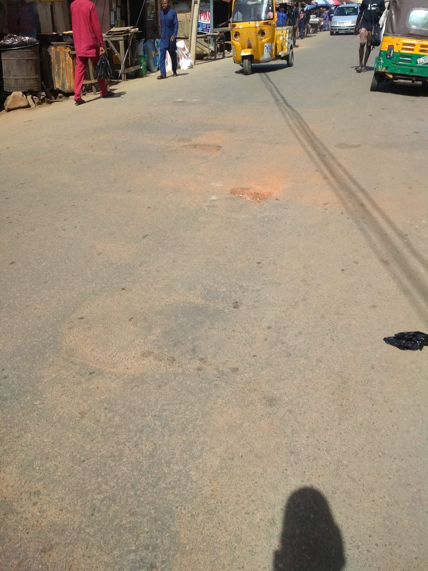 This image depicts a bustling street scene in Jos, Nigeria, bathed in strong midday sunlight. The asphalt road surface shows signs of wear, including several small reddish-brown depressions and long, faint tire marks extending diagonally across the center of the frame.
Several people are visible. In the foreground on the left, a man dressed in a bright red traditional Nigerian tunic and trousers, carrying a black bag, walks away from the camera. Further back on the left, another man in a blue traditional outfit walks, looking towards the right. On the far right, a person in a dark t-shirt and shorts, also carrying a bag, walks away. The shadow of the person taking the photograph is visible in the bottom right corner of the frame.
Vehicles include a yellow three-wheeled auto-rickshaw (Keke Napep) with the number "7109" on its front, positioned in the midground. The rear of a green and yellow Keke Napep, displaying a partial license plate "SSA-964", is visible on the far right. A silver car can be seen in the distant background.
The left side of the street features various commercial elements such as barrels, wooden stalls, and sacks, suggesting a market or street vendor area. A "POS" (Point of Sale) sign is discernible behind the man in blue. The strong shadows cast by people and vehicles indicate a bright, sunny day. A crumpled black plastic bag lies on the road surface towards the bottom right.