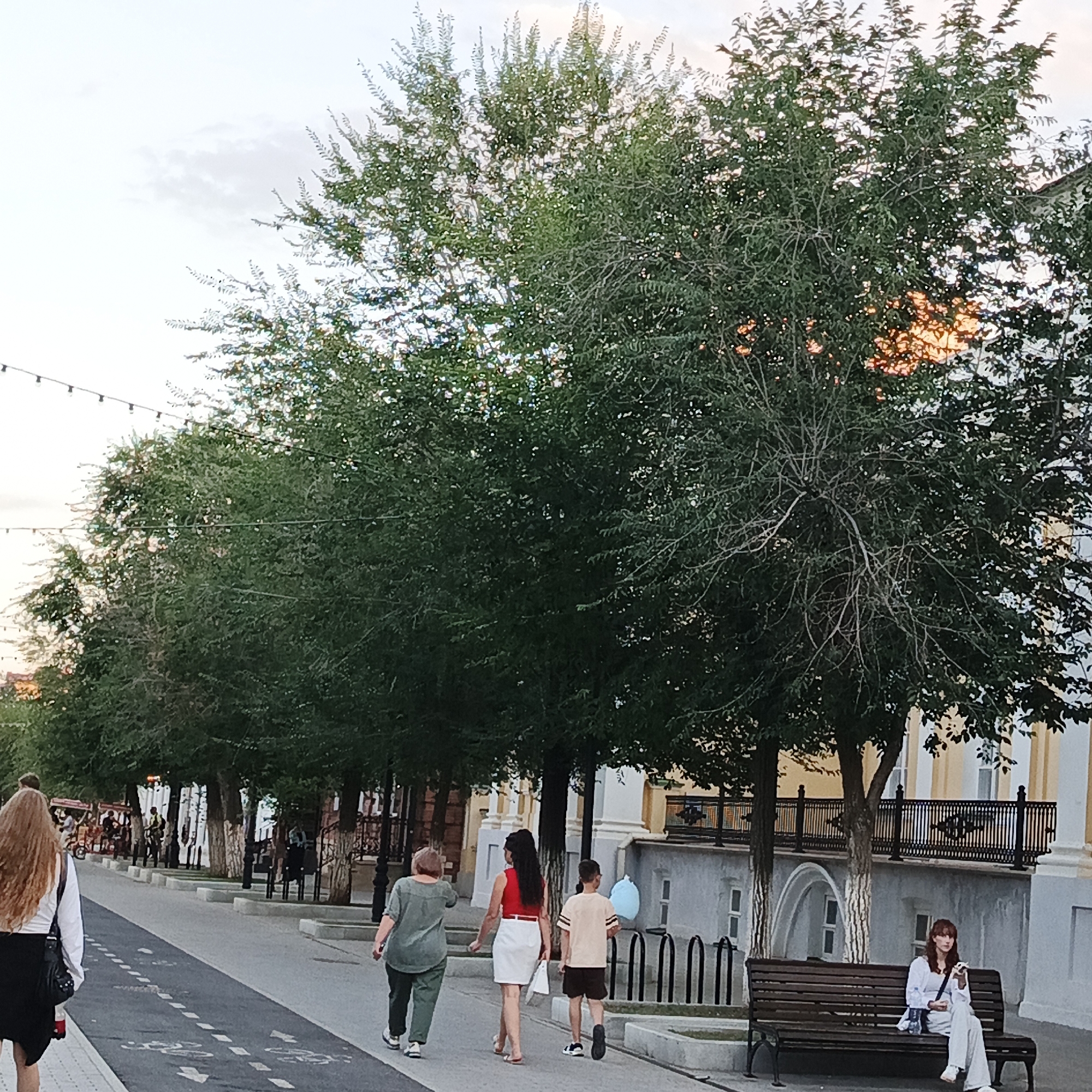The image shows a street scene in Orenburg, Russia, likely on Sovetskaya Street given the pedestrian and cycling paths, and the buildings lining the street. In the foreground, a woman with long, wavy brown hair walks away from the camera on a pedestrian path marked with dashed lines and bicycle symbols. She is wearing a white long-sleeved shirt and a black skirt, carrying a black purse. To her right, on the same path, a person wearing a green shirt and dark pants walks in the same direction. Further ahead, a woman in a red top and white skirt walks, accompanied by a boy in a beige t-shirt and brown shorts. They are walking on a paved path next to a row of trees. On the right side of the image, a woman with reddish hair sits on a wooden bench beneath a tree, holding a white object, possibly a phone or food. Behind her, a building with yellow and white accents and black railings is visible. The street is lined with trees, and there are streetlights in the distance. The sky is overcast with hints of sunlight breaking through.