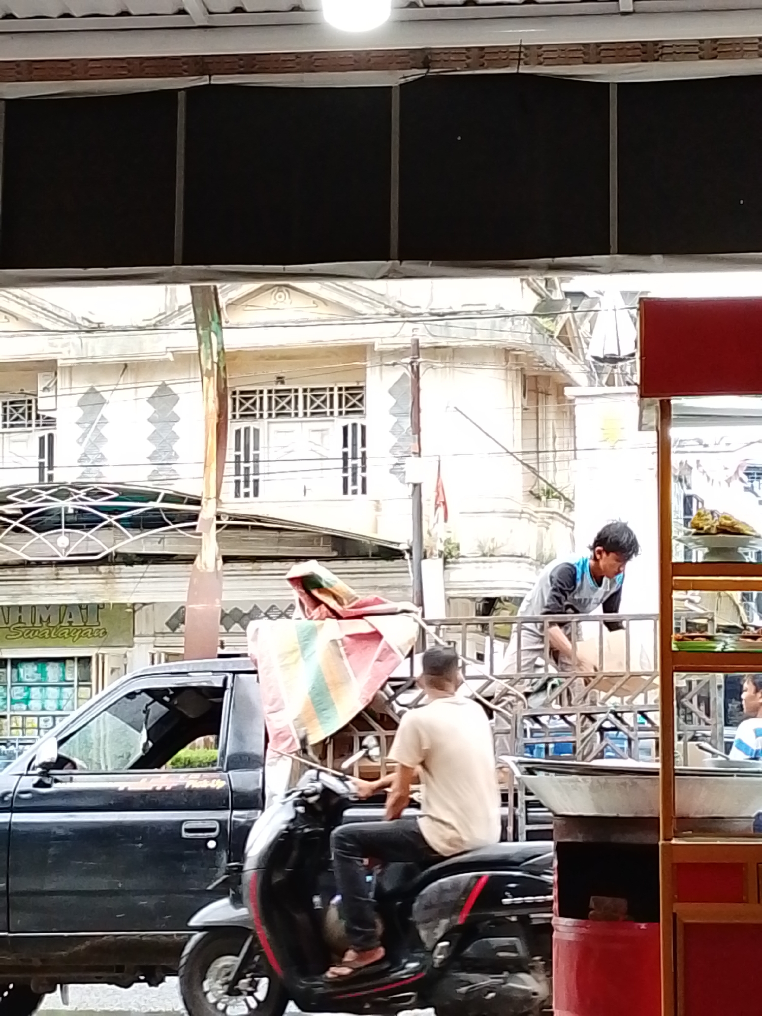 The image captures a bustling street scene in Lhokseumawe, Indonesia, during daytime. In the foreground, a man wearing a beige shirt and dark pants rides a black scooter, facing away from the viewer. To his left, a dark-colored pickup truck is seen in profile, partially obscured. On the truck's bed, a colorful striped tarp or umbrella is draped.
Further into the midground, a young man in a grey and blue t-shirt and light-colored pants is standing in the bed of what appears to be another truck or a cart attached to a vehicle, possibly arranging goods or working. To the far right, a red-framed street food stall is visible, displaying various food items, including some yellowish/greenish dishes on tiered trays and large metal cooking pots.
The background features several light-colored buildings with ornate architectural details, including decorative window grilles and molding, suggesting a mix of styles, possibly colonial or traditional Indonesian. Partially visible on one building is a sign that reads "HMAT Swalayan," indicating a supermarket or convenience store. The sky is not visible, but the bright lighting suggests it is sunny. The overall scene depicts everyday life and commerce in an urban setting.