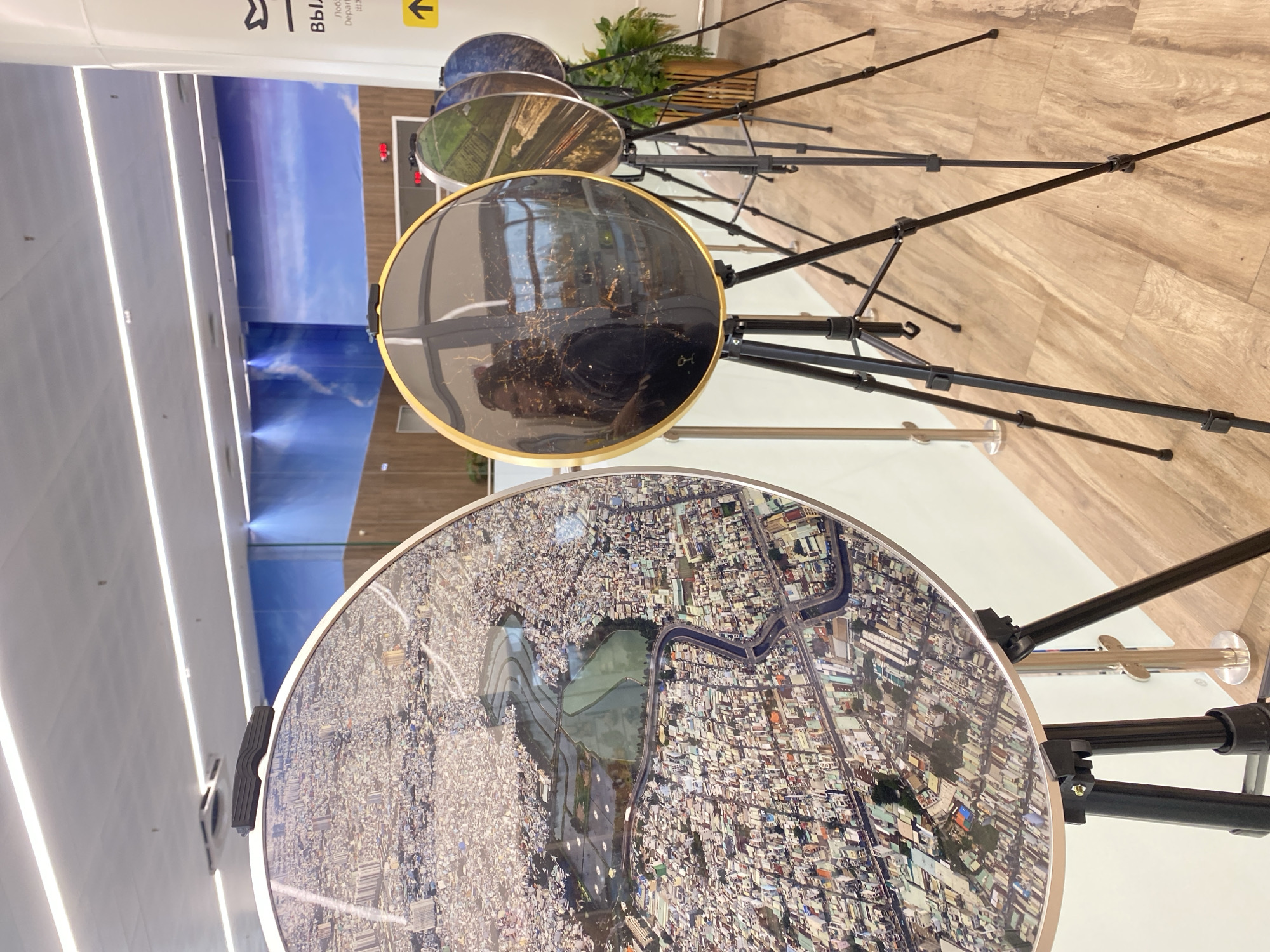 The image shows a display of four circular artworks, each mounted on a tripod. The first artwork on the left is a detailed aerial view of a densely populated urban area with a winding river and green spaces. The second artwork, in the center, is a darker image, possibly a nighttime cityscape or reflection, framed by a gold border. The third and fourth artworks, to the right, show aerial views of green fields and what appear to be more distant urban areas, with the fourth artwork being partially obscured. The artworks are displayed in an indoor environment with a light-colored floor and a backdrop that appears to be a mural of a blue sky with clouds. There are also signs visible on a white pillar to the right, one with a yellow arrow pointing upwards and text in Russian and what appears to be Chinese characters, indicating "Departure". The scene is well-lit by overhead fluorescent lights. There are no people clearly visible within the scene. The location of Новое Мартьяново, Russia is not ascertainable from the visual information provided in the image.