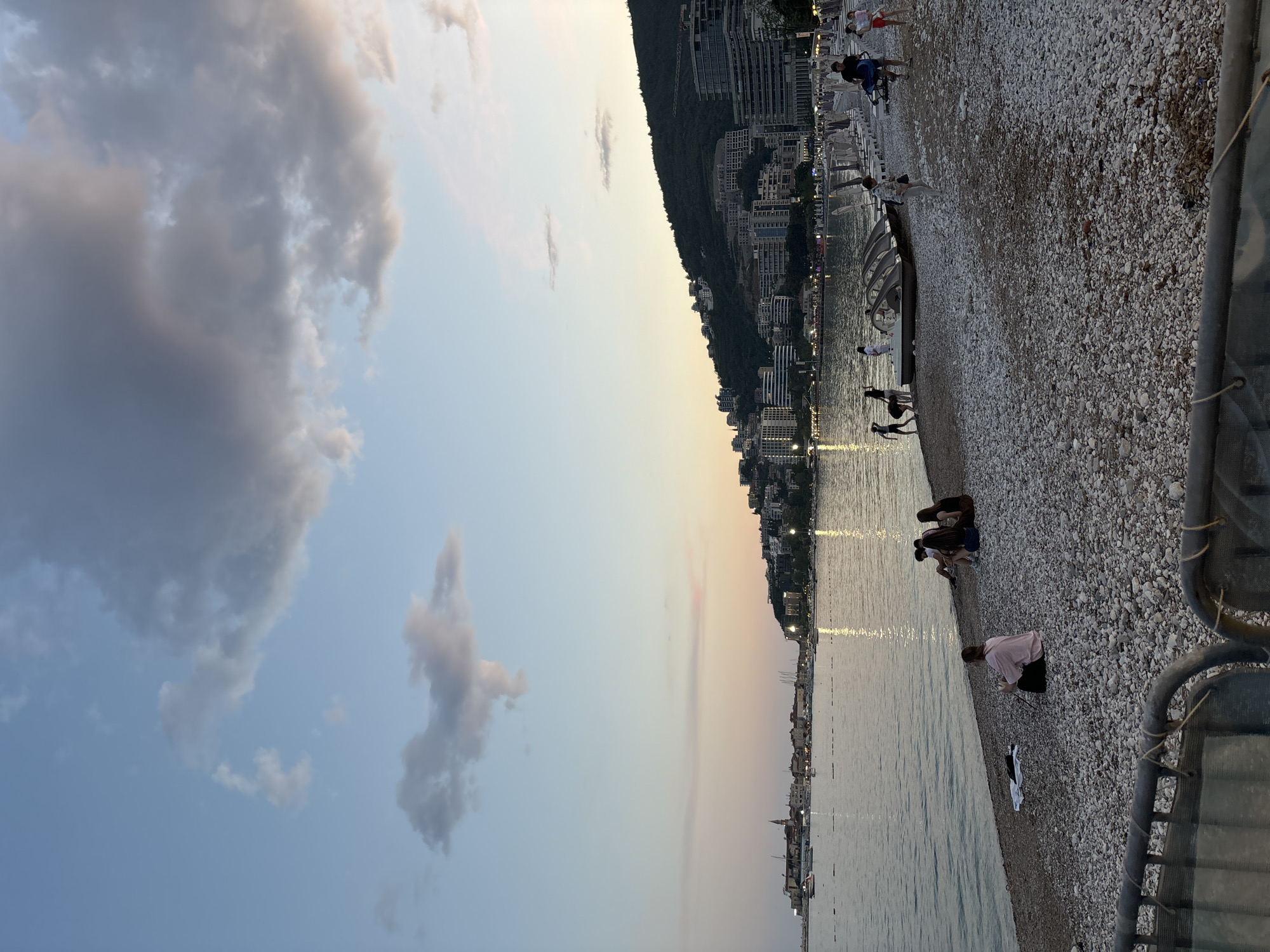 The image shows a pebble beach in Budva, Montenegro at dusk. People are walking or sitting on the beach. Buildings line the coast behind the beach. Clouds are visible in the sky. The water is calm. A fence borders the beach on the right side of the image.
