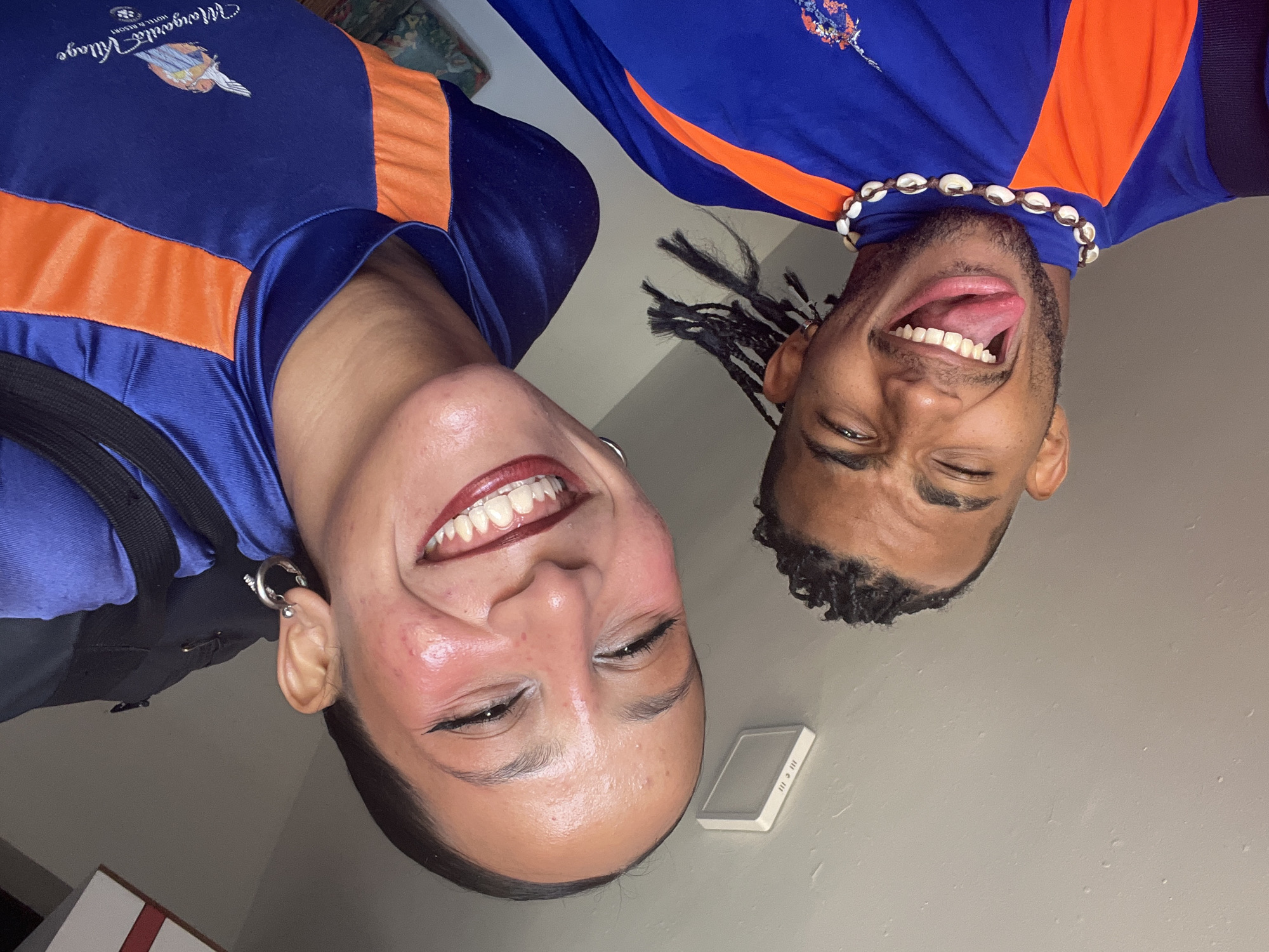 A selfie features two individuals wearing blue and orange collared shirts, likely taken indoors against a light-colored wall. The individual on the left is smiling widely, displaying braces. The individual on the right is sticking out their tongue and wearing a necklace of white shells. A small white object is on the wall near the smiling person, and a black bag strap is visible on the left. The shirt on the left appears to have the logo "Margaritaville Village." The exact location within the city of Porlamar, Venezuela, cannot be determined from the visual information provided.