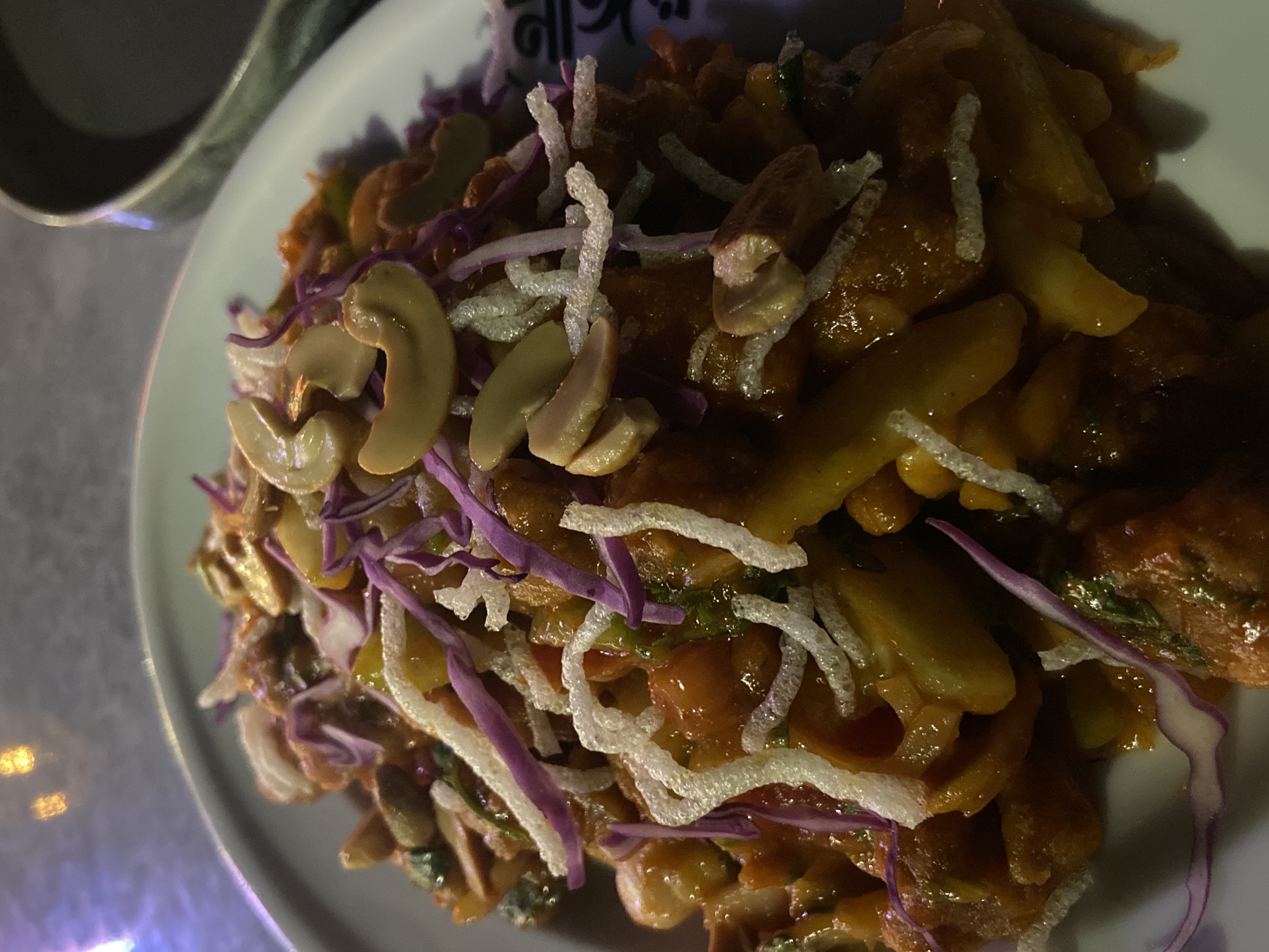 A plate of food is presented on a table, likely photographed indoors in Dhaka, Bangladesh. The dish features stir-fried potatoes coated in a brown sauce, topped with garnishes of shredded red cabbage, white puffed rice noodles, and cashews. A container sits near the plate. The table surface is textured and has a light source creating a reflection.