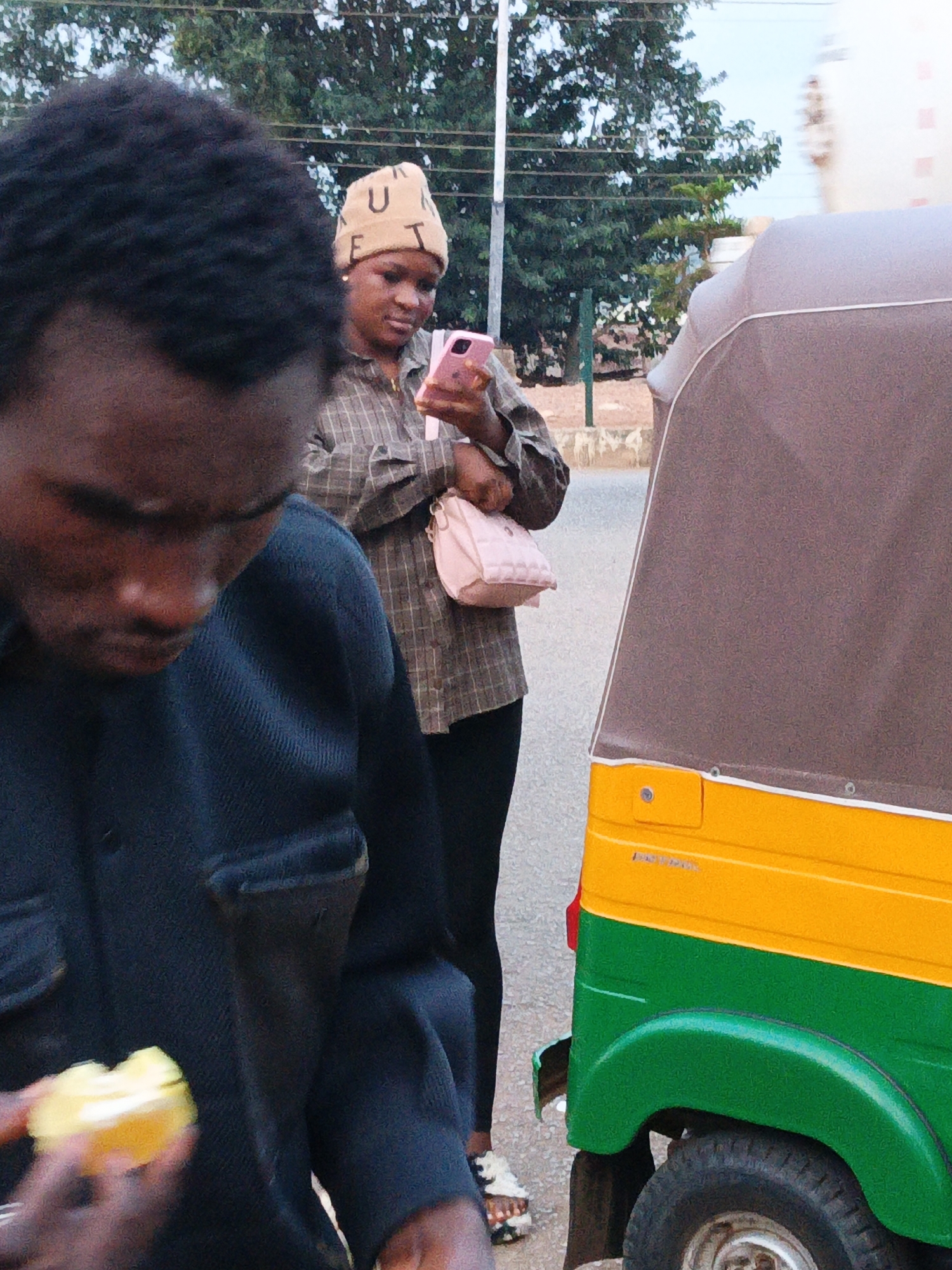 In an outdoor urban scene, a person in the foreground with dark, textured hair is partially visible, looking down and holding a yellow object in their hand, possibly food. Behind them, a woman wearing a light-colored beanie with letters on it, a checkered long-sleeved shirt, and dark leggings is focused on her pink smartphone. She holds a small, quilted light pink purse in her left arm. To the right, a portion of a colorful vehicle, possibly an auto-rickshaw, is visible. The vehicle has a brown canopy over a bright yellow and green body. The background consists of trees, vegetation, and a light-colored sky. The ground appears to be paved.