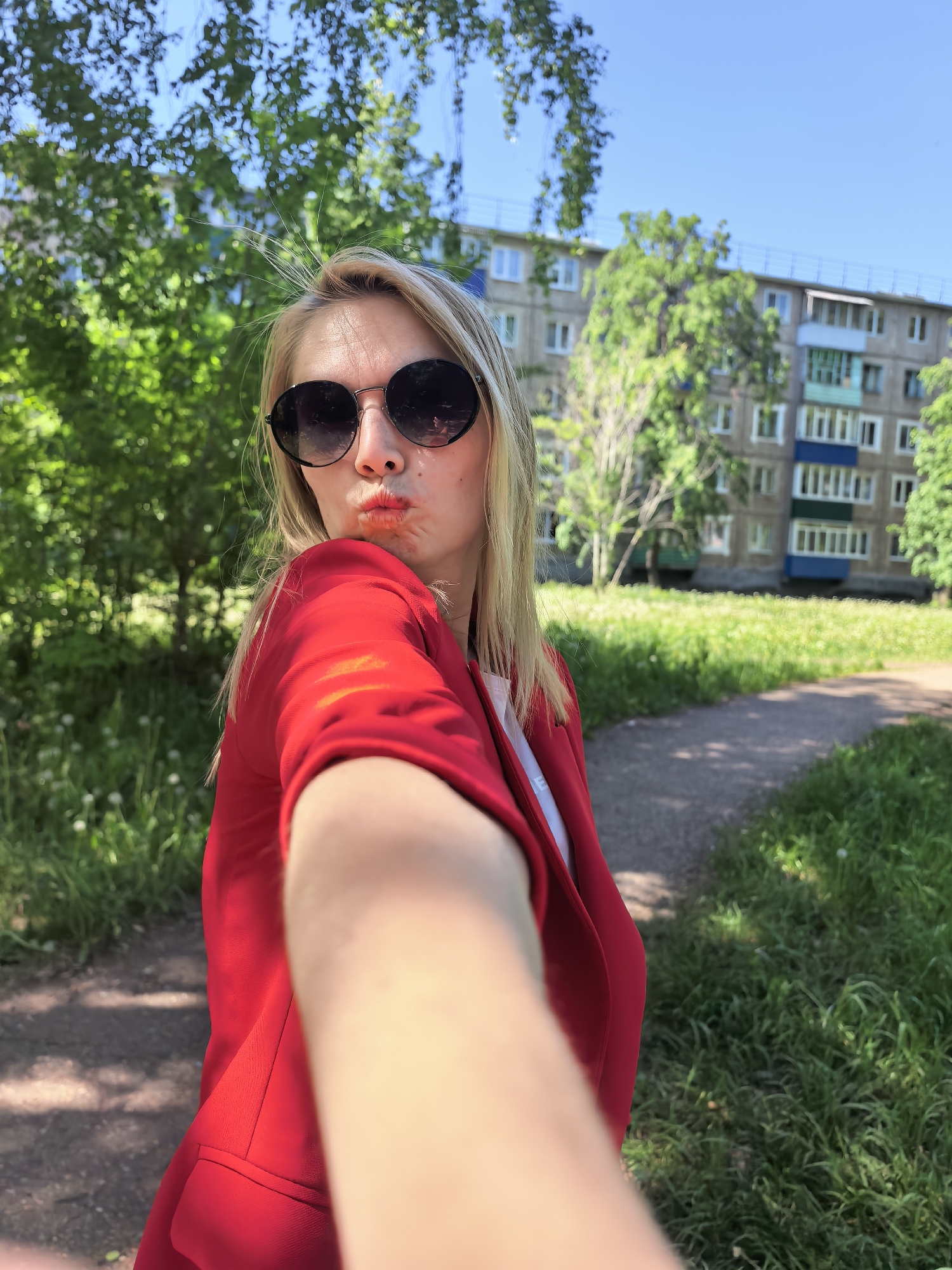 A fair-skinned woman with blonde hair, wearing round sunglasses and a red blazer, is taking a selfie outdoors. She is positioned in front of a tree-lined pathway and building. The location is Sterlitamak, Russia.