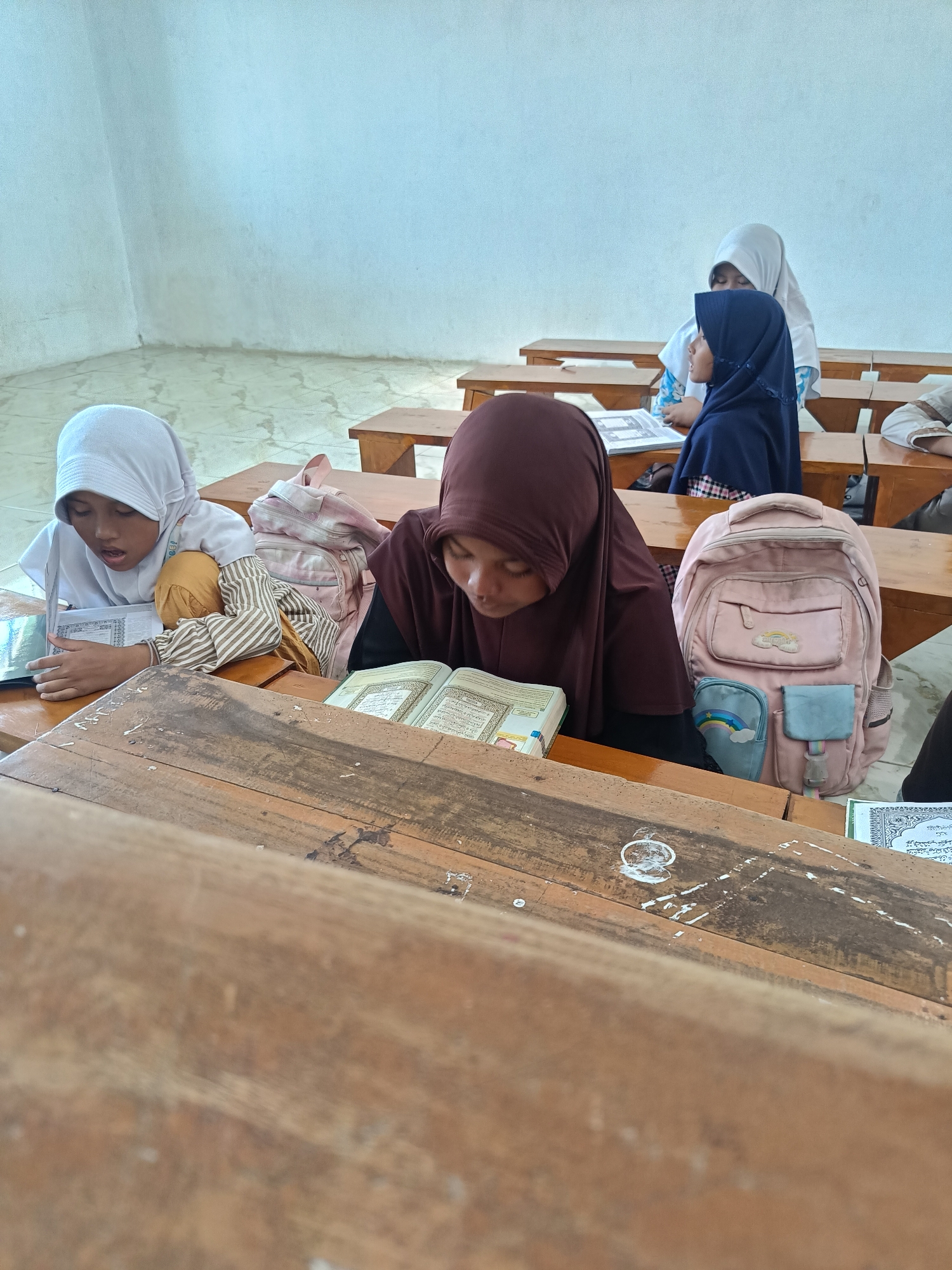 In a classroom in Pakijangan, Indonesia, four young girls wearing hijabs are seated at wooden desks, engrossed in reading. The girl in the foreground on the left wears a white hijab and is actively reading from an open book placed on her desk. Beside her, a pink backpack with cloud and rainbow patches rests on the desk. Another girl, closer to the center, is also reading from an open book on her desk, her dark maroon hijab obscuring her face as she focuses intently. In the background, two other girls are visible, one with a white hijab and the other in a dark blue hijab, also engaged in reading. The classroom has light blue walls and light-colored tiled flooring. Sunlight streams in from the left, illuminating the scene and casting soft shadows. The wooden desks are worn, suggesting regular use.
