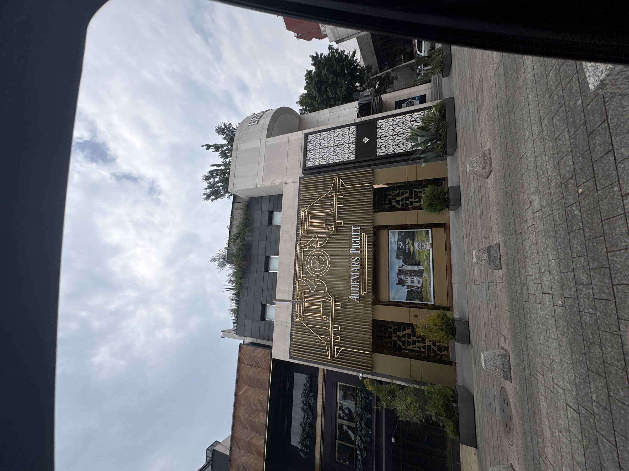 The image shows the Audemars Piguet store in Mexico City, Mexico. The building is multi-storied with a light-colored facade and a prominent gold-toned logo displaying the brand name. The storefront includes a window display. The building has decorative elements, including a patterned black screen-like structure above the logo. Plants are incorporated into the building's design. The sidewalk in front of the store is paved with grey stones. The image appears to have been taken from inside a vehicle. The sky is overcast.