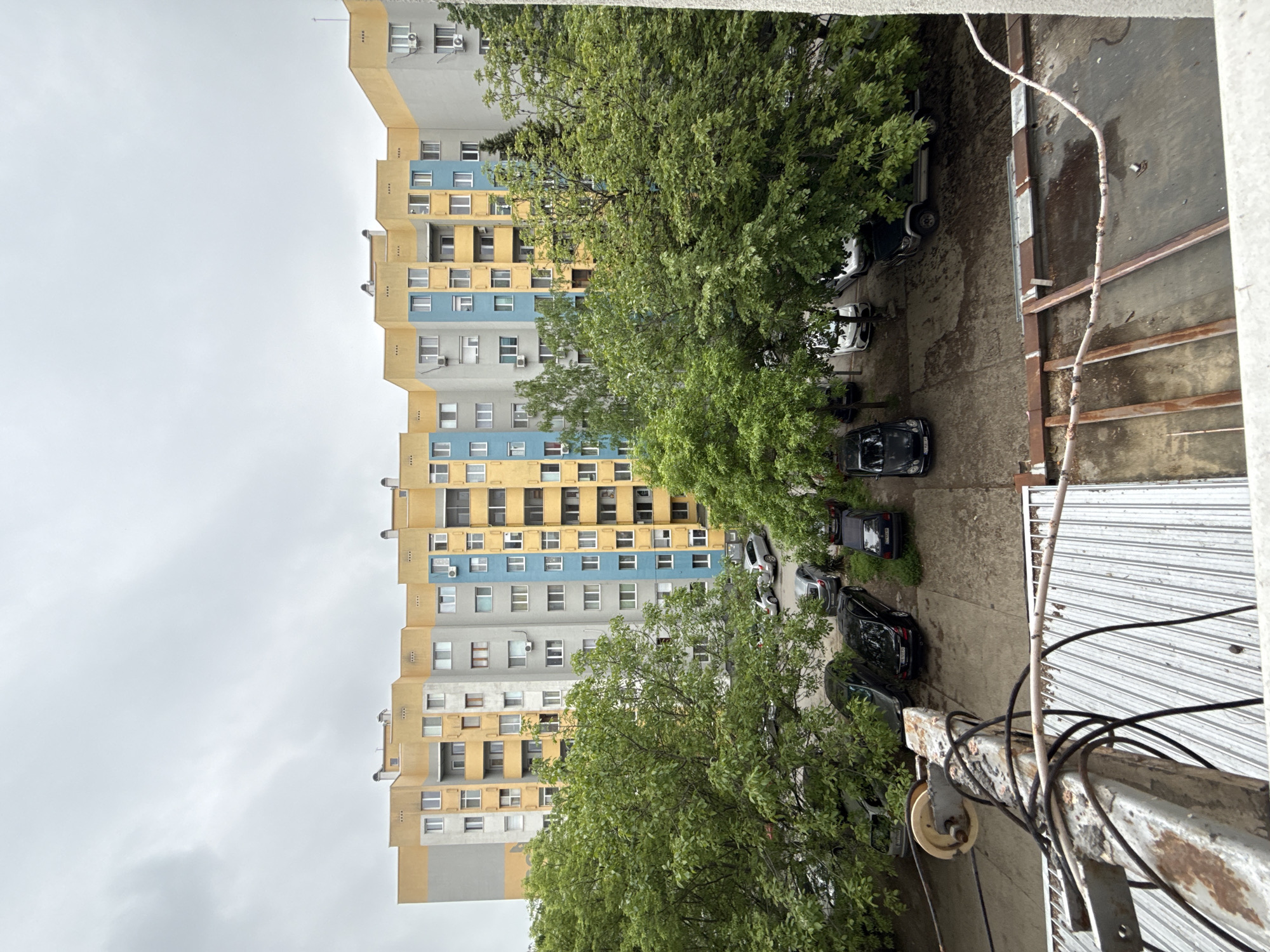An upward-angle shot shows a tall residential building in Stara Zagora, Bulgaria, partially obscured by lush green trees. The building features a repetitive pattern of grey, yellow, and blue panels, punctuated by rows of windows, and some with visible air conditioning units. The building is centered on the left side of the frame. Beneath the trees is a concrete paved area with parked cars, including white, black, and grey vehicles. On the right side of the frame is a close-up view of an adjacent metallic balcony structure with visible wear and tear. Several black cables are attached to it. The sky above is overcast with grey clouds.