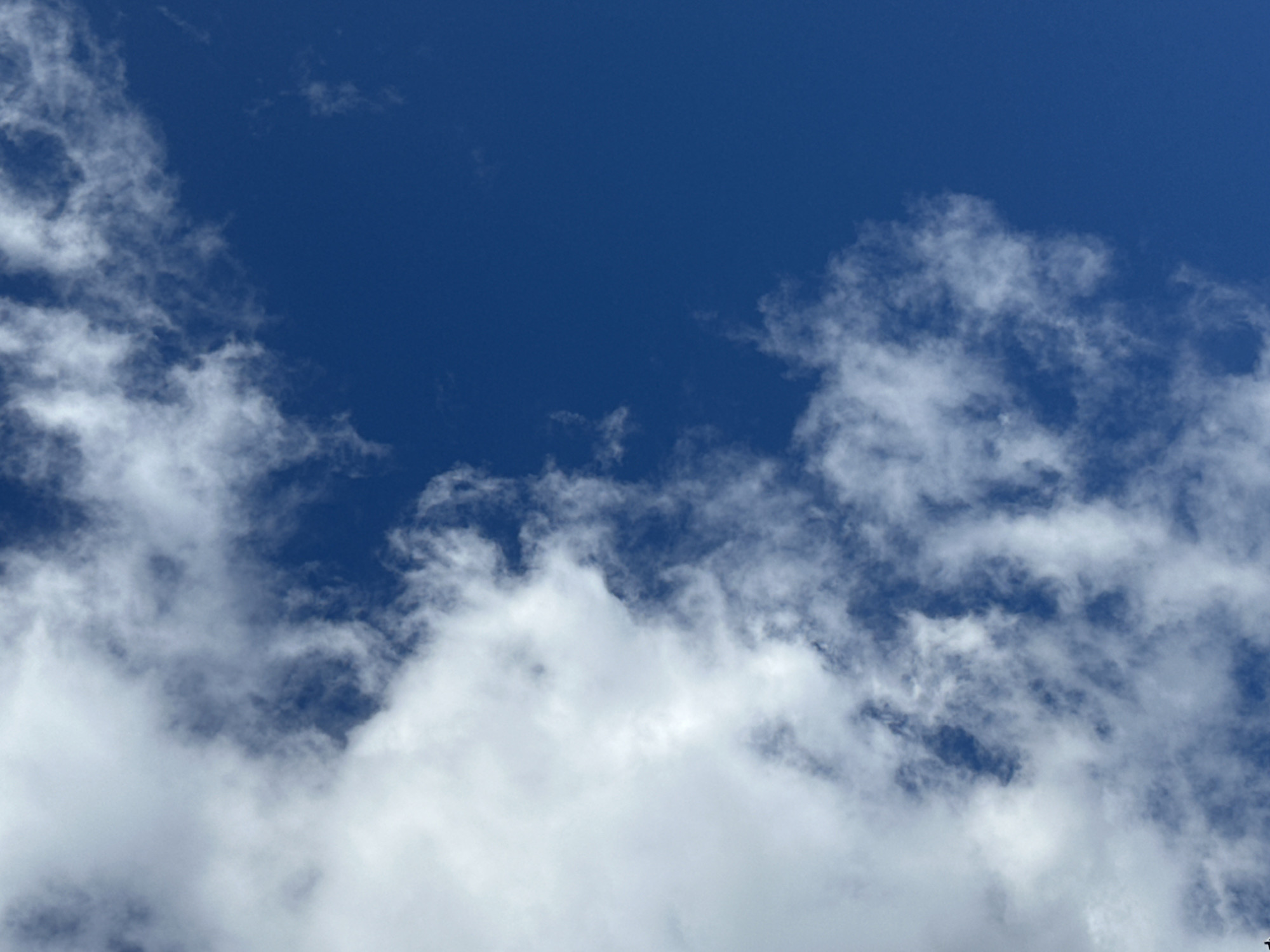 The image captures a bright blue sky with scattered white clouds. The clouds vary in density, with some appearing wispy and thin, while others are more substantial and billowy. The sky takes up the majority of the frame, with clouds occupying the left and upper portions, thinning out towards the right side. The image appears to be an outdoor scene, likely taken during the daytime, given the brightness of the sky. No specific location within a city, such as Unknown, Russia, can be identified from the image content. There are no discernible people, subjects, objects, actions, or interactions depicted.
