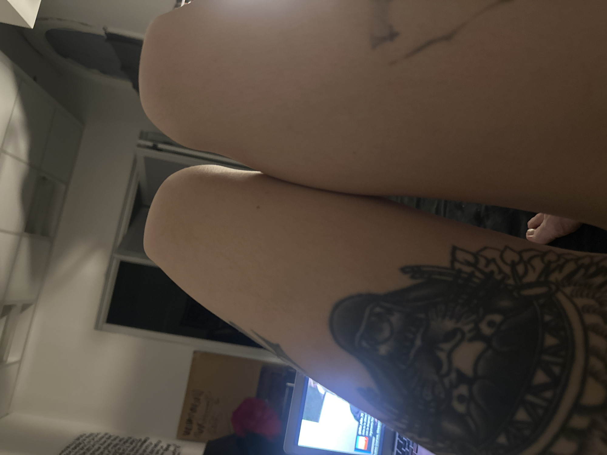 A close-up, tilted shot depicts a person's thighs with tattoos. The upper thigh has a faint, undefined tattoo. The lower thigh features a detailed tattoo of a stylized image. A laptop screen is visible below the lower thigh, suggesting the person is likely seated indoors. The environment includes a white wall with tiled panels, a window, and a cardboard box in the background. There is no verifiable evidence to indicate the location is Buenos Aires, Argentina.