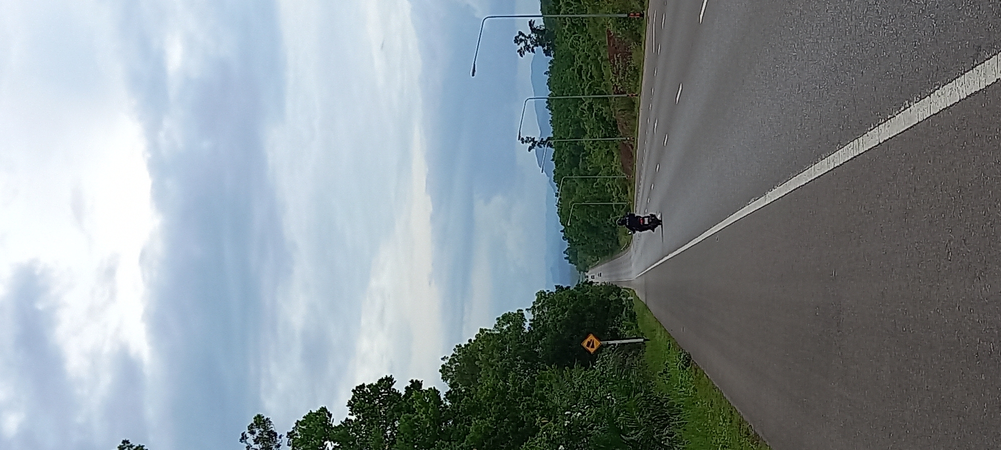 The image depicts a long, straight asphalt road in Khian Sa, Thailand, with a white line marking the edge. A motorcycle is traveling along the road. The environment includes trees and vegetation alongside the road. Streetlights are positioned at regular intervals. The sky is overcast. A road sign is visible on the roadside.