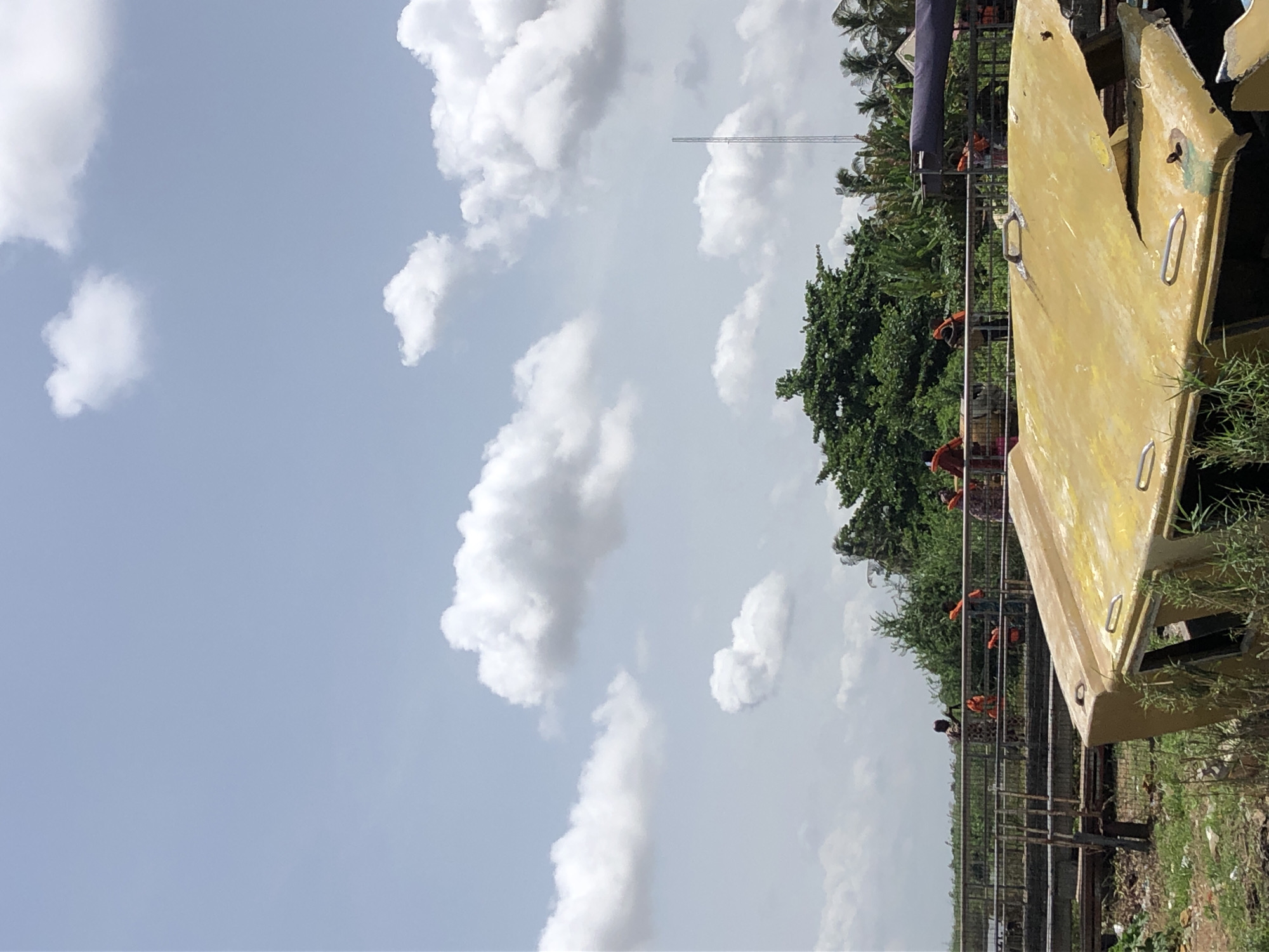 The image displays an outdoor daytime scene under a bright, partly cloudy sky. In the foreground to the right, a large, yellow, boat-like structure or hull rests on overgrown ground, surrounded by grass and some debris. It has several metal handles or cleats on its side.
Extending horizontally in the mid-ground is a metal walkway or bridge, upon which several people are visible. Some of these individuals are wearing bright orange life jackets. To the left of the walkway and stretching towards the background is a dense canopy of lush green trees. A tall, slender mast or antenna stands in the distant background, rising above the treeline. A dark blue or purple tarp or fabric is partially visible in the upper right portion of the image.
The scene suggests a location near water, such as a riverbank or lake, possibly in a tropical or subtropical region, given the vegetation and the presence of life jackets. The time of day is clearly daytime with bright sunlight, and the weather appears fair with scattered cumulus clouds. No visible text is present in the image.