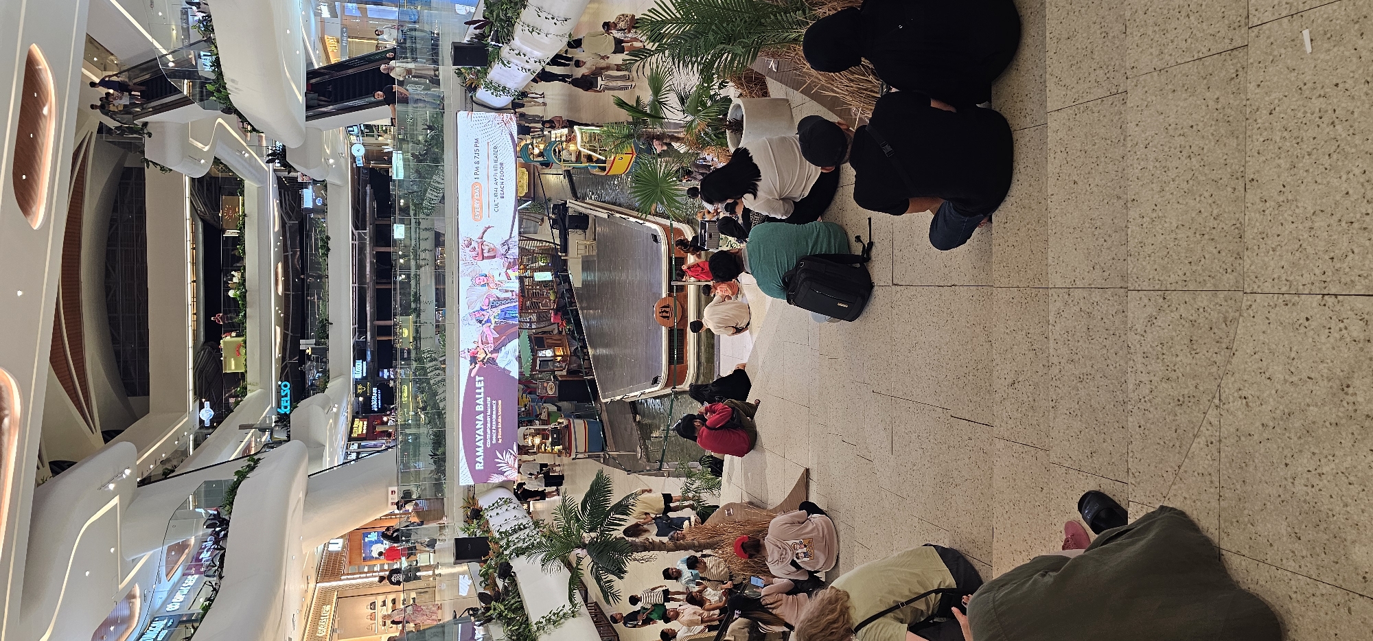 The image depicts an event in what appears to be a multi-level shopping mall. A crowd of people is gathered on a light-colored, tiled floor, seemingly watching a performance on a boat, which is positioned in front of a large screen displaying "RAMAYANA BALLET". Palm trees and other greenery are placed around the performance area. The architecture of the mall features curved, modern designs, with shops visible on multiple floors. The location is likely the city of Denpasar, Indonesia. The event seems to be well-attended, with people sitting or standing to observe the performance.