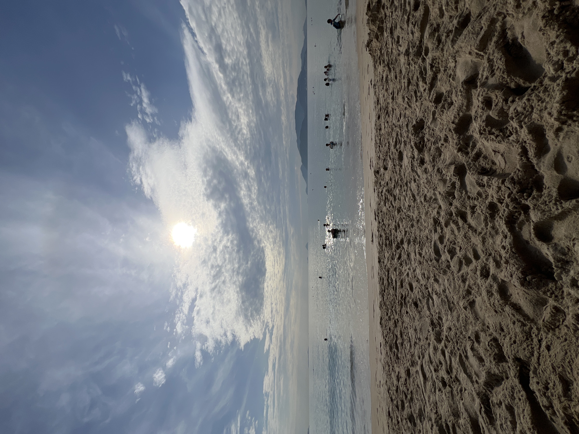 The image depicts a beach scene in Nha Trang, Vietnam, likely during daytime. The foreground is dominated by a sandy beach with footprints visible. The middle ground features the ocean with several people swimming and playing in the water. Some individuals are partially submerged, while others appear to be standing. The background shows a calm sea extending to the horizon, with a hazy mountain range visible in the distance. The sky is partly cloudy with the sun shining brightly, creating sunbeams through the clouds. The overall atmosphere is one of leisure and recreation.